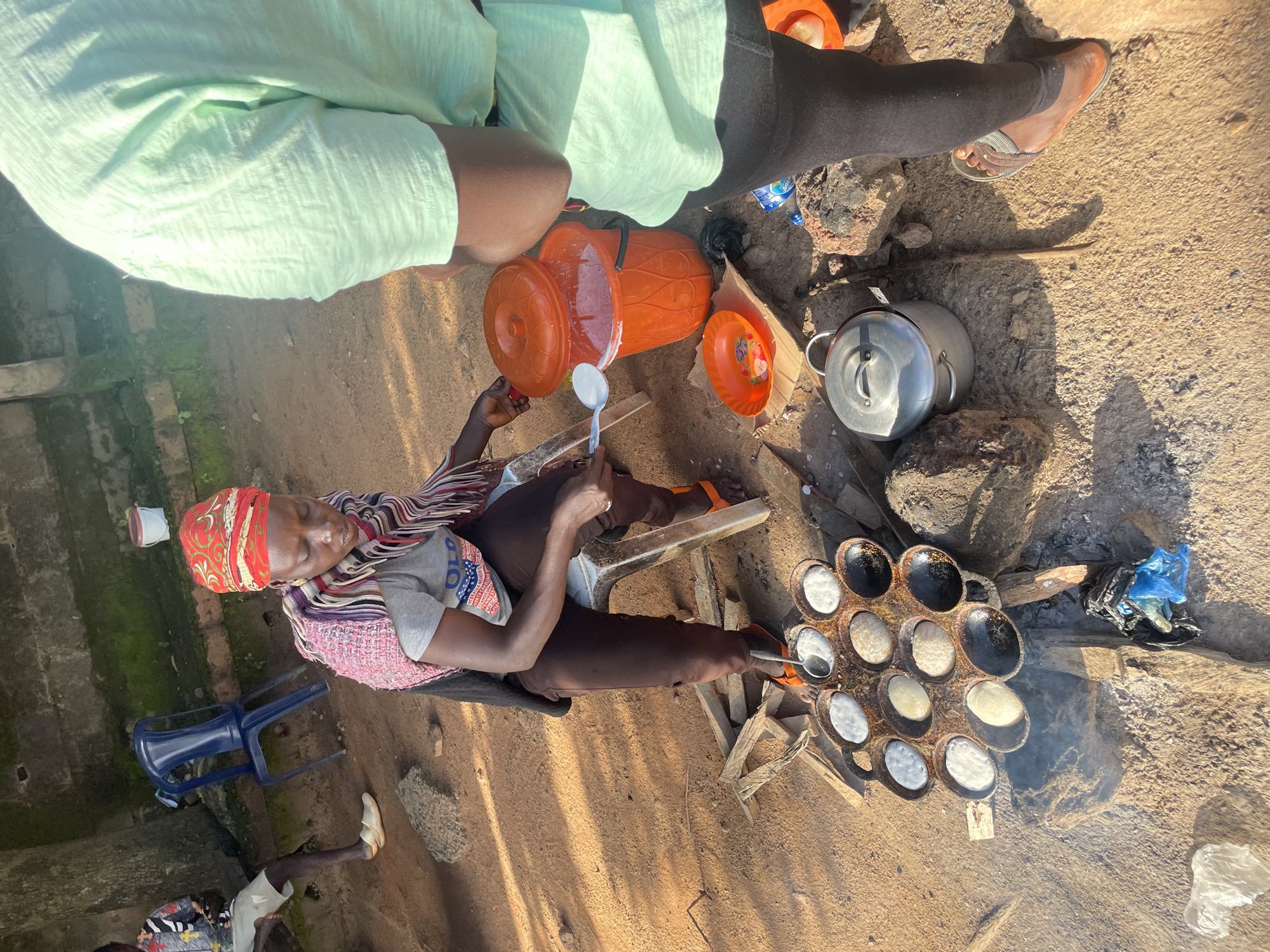The image depicts an outdoor cooking scene in the city of Jos, Nigeria. A woman is seated on a stool, wearing a red headscarf and a striped scarf draped over her shoulders. She is actively cooking, using a spoon to add batter to a traditional cooking pan placed over a fire. The pan has multiple circular indentations, some filled with white batter, others with cooked food, and some appearing burnt. Next to her, an orange bucket with a lid is filled with a white liquid, likely batter. A small orange plate with a pattern is also nearby. In the foreground, a large metal pot with a lid rests on the ground. Another person, partially visible, is standing in the right foreground, wearing a light green shirt and black leggings. In the background, a blue plastic chair and a wall with moss growth are visible, suggesting an informal outdoor setting. The ground is covered in dirt and scattered debris.