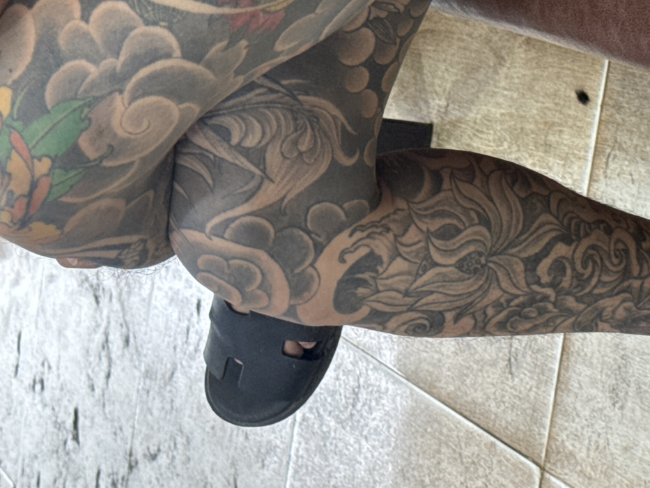 A close-up, downward-angled view captures the lower body of an individual seated in Nha Trang, Vietnam. The right leg is crossed over the left. Both visible legs are extensively covered in traditional Japanese-style tattoos, predominantly in grey wash, depicting swirling cloud/wave patterns and detailed lotus flowers. Minor accents of red, green, and yellow are visible on the upper right portion of the right leg. The left foot wears a black, two-strap slip-on sandal. The subject is positioned on a floor composed of light grey or beige rectangular tiles with a textured surface. A dark vertical object and a light wooden surface are partially visible in the background to the right.