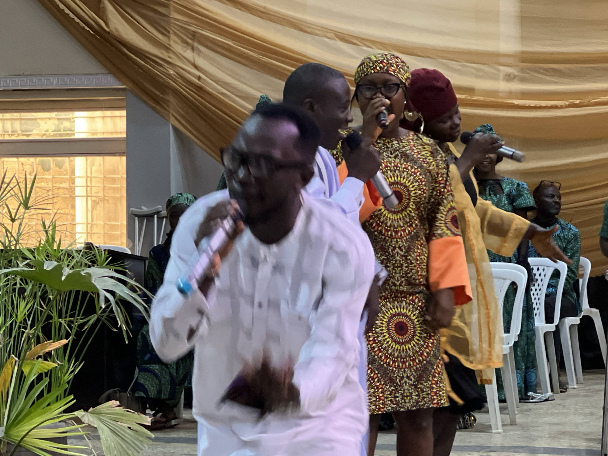 In Kabong, Nigeria, a group of people are gathered indoors, possibly for a cultural event or performance. The setting appears to be a hall, with a backdrop of draped golden fabric. In the foreground, a man in a white garment is actively singing into a microphone, his body in motion. He is surrounded by other individuals, some of whom are also holding microphones and singing. One woman in the foreground wears a vibrantly patterned dress and a matching headscarf, singing with intensity. To her right, another woman in a maroon headscarf also holds a microphone. The individuals are positioned closely together, suggesting a collaborative performance. In the background, several white chairs are arranged in a row, with people seated and observing. The overall atmosphere seems lively and engaged, with a focus on music and vocal performance. The presence of microphones and the singing action strongly indicate a musical or spoken word performance is taking place.