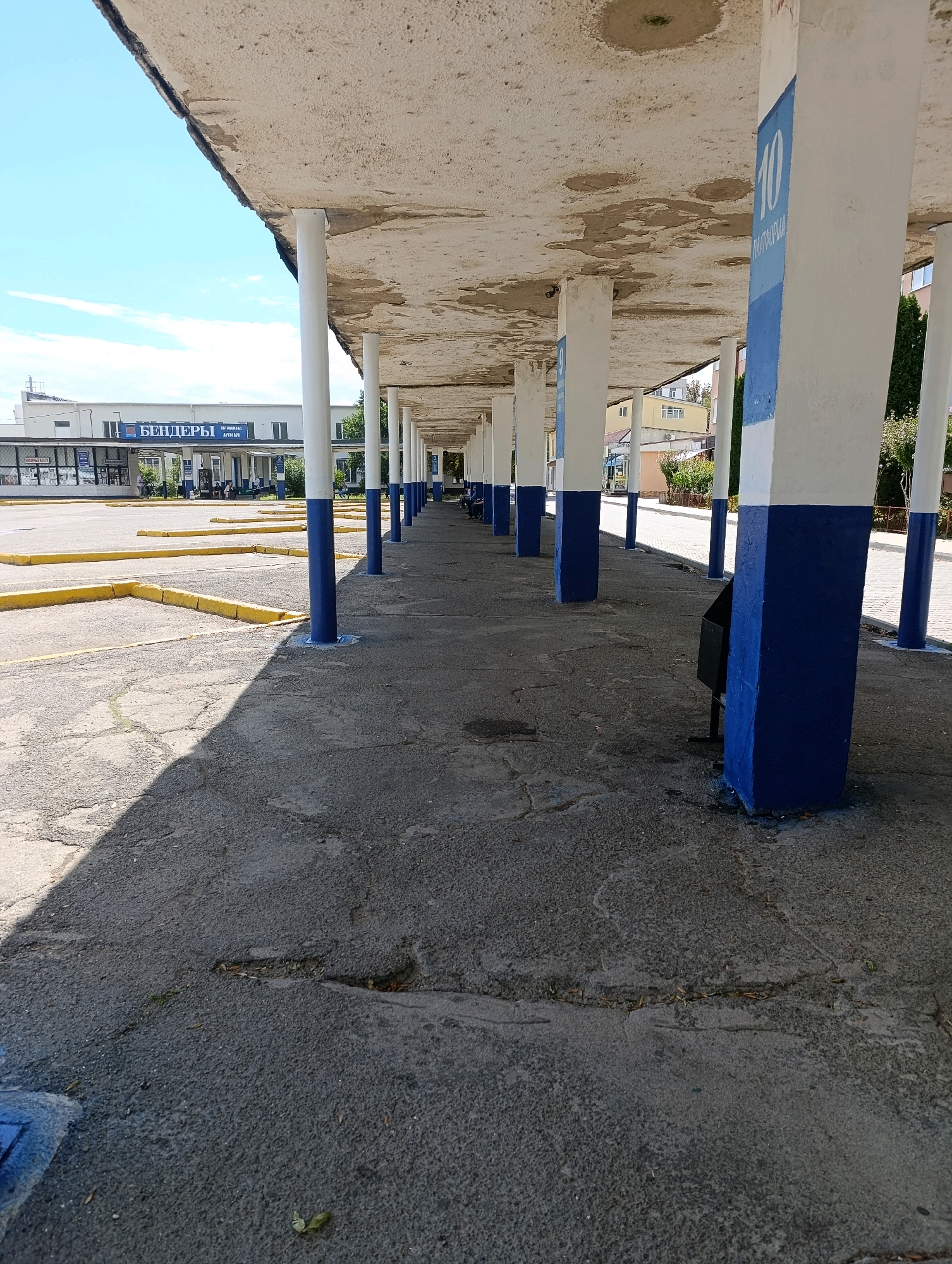 The image shows a covered walkway or bus stop area in Bender, Moldova. The walkway is supported by a series of white columns painted with blue bases, and there are blue and white signs on some of the columns, one of which clearly displays the number "10". The roof of the walkway is made of concrete and appears to be weathered and in some areas, damaged. The ground surface is paved with asphalt and shows signs of wear and cracking. In the background, there is a building with signage in Cyrillic, which reads "БЕНДЕРЫ" (Benders). Yellow lines on the asphalt in the foreground suggest designated parking or waiting areas. The scene is outdoors, under a bright sky with some clouds. No individuals are clearly visible or engaged in any specific actions.