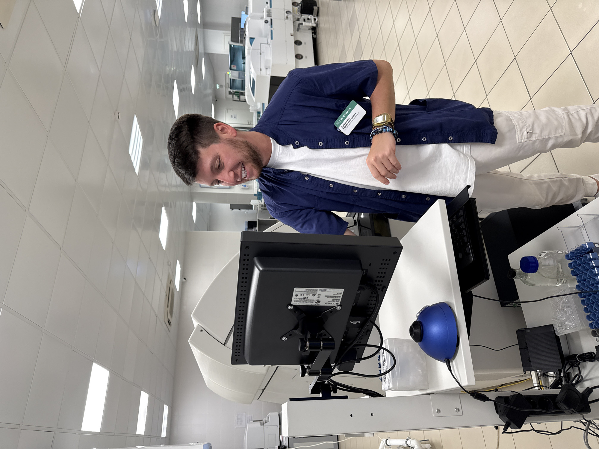 A young man with short brown hair and a beard stands indoors, smiling and looking down. He is wearing a white t-shirt, a blue short-sleeved button-down shirt, and white pants. A name tag is visible on his shirt.
In front of him is a computer monitor and associated equipment on a white countertop. A blue object is also on the counter.
The background shows what appears to be a laboratory or medical facility with white walls, tiled floors, and various pieces of equipment. The setting could possibly be in the city of Moscow, Russia, though this is primarily indicated only from text on the name tag.