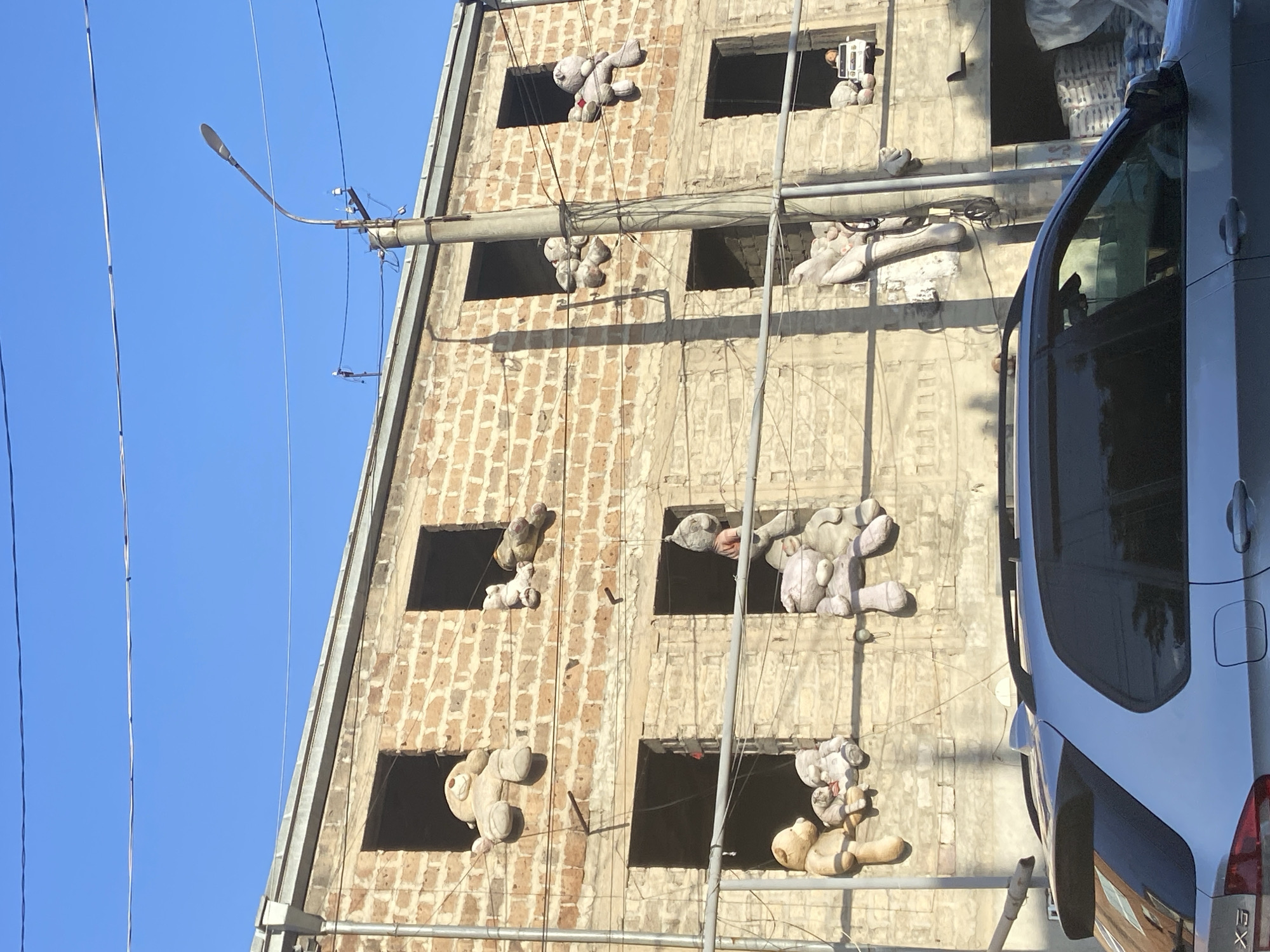 The image shows the exterior of a multi-story building in Yerevan, Armenia, under a clear blue sky. The building appears to be under construction or renovation, with empty window frames. Several large, stuffed teddy bears are positioned in the window openings, some hanging by what appear to be wires or ropes. A street light and power lines are visible in the foreground. A silver van is parked on the right side of the frame, partially obscuring the building. The building's facade is made of brick.