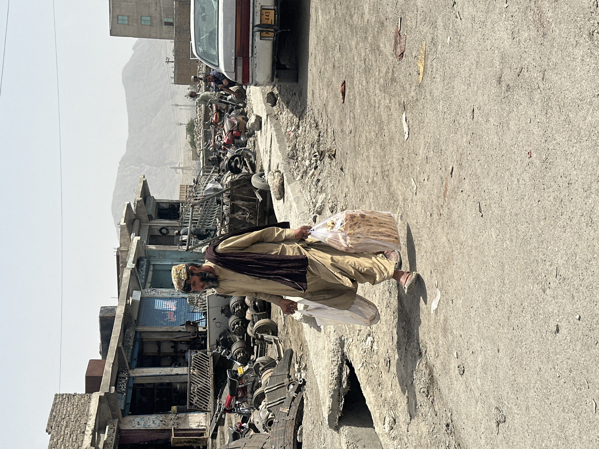 A man in traditional Pakistani clothing is walking down a street in Quetta City Tehsil, Pakistan. He is carrying two plastic bags filled with bread. He has a brown shawl and a patterned hat. The street is cluttered with motorcycle and car parts. There are also several buildings, and a white truck is partially visible in the background. A mountain range is visible in the background under a bright sky.