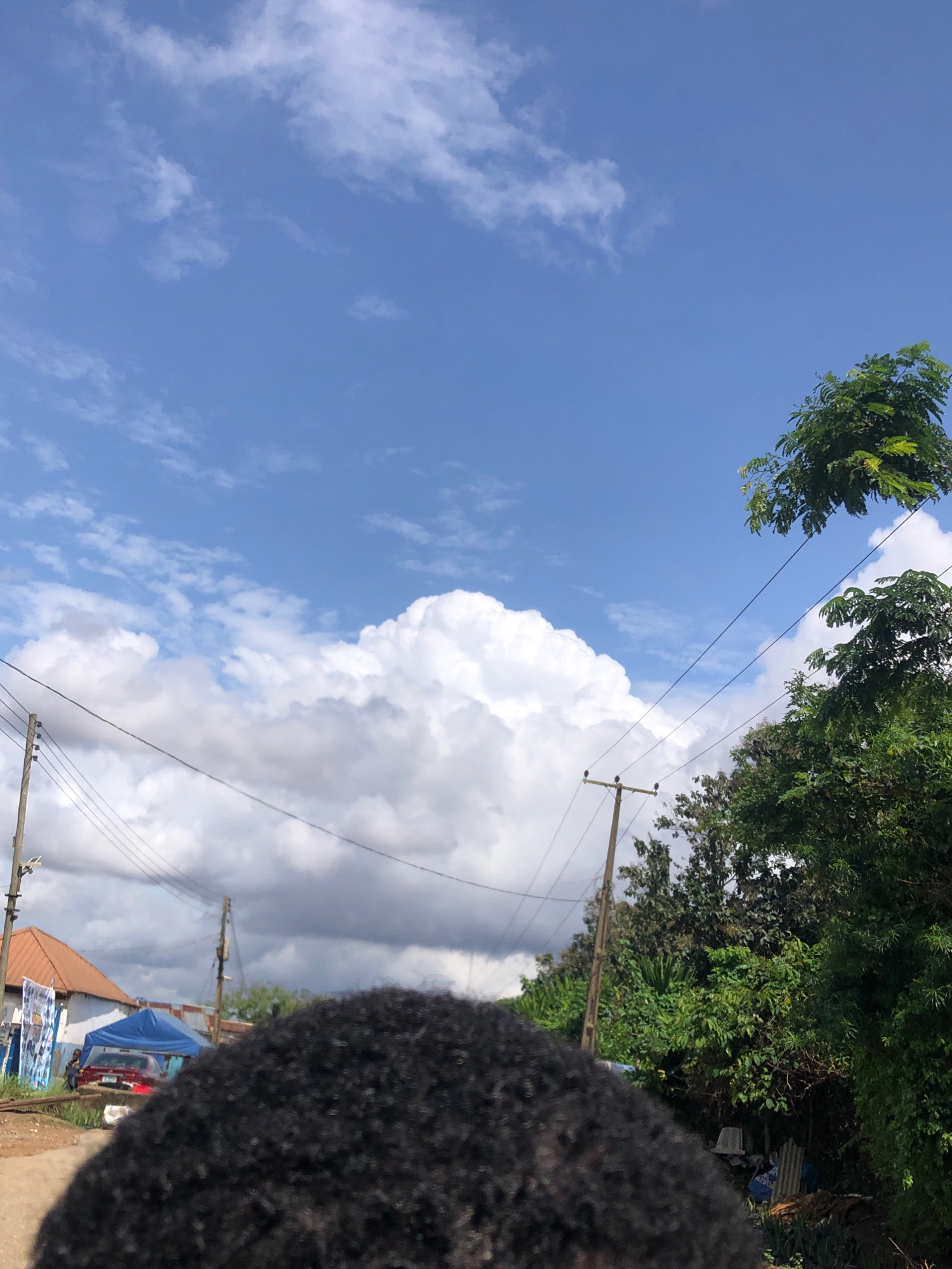 In the city of Agege, Nigeria, a person's dark, curly hair is in the foreground of the image, obscuring much of the lower half of the frame. The background reveals a street scene with a clear blue sky dotted with white clouds. Several utility poles with wires traverse the sky, connecting to unseen structures. On the left side of the image, a building with a reddish-brown roof and some signs is visible, along with a red car parked under a blue tent. Dense green foliage and trees are present on the right side, partially obscuring other structures and objects. The overall impression is of an outdoor setting on a bright day.