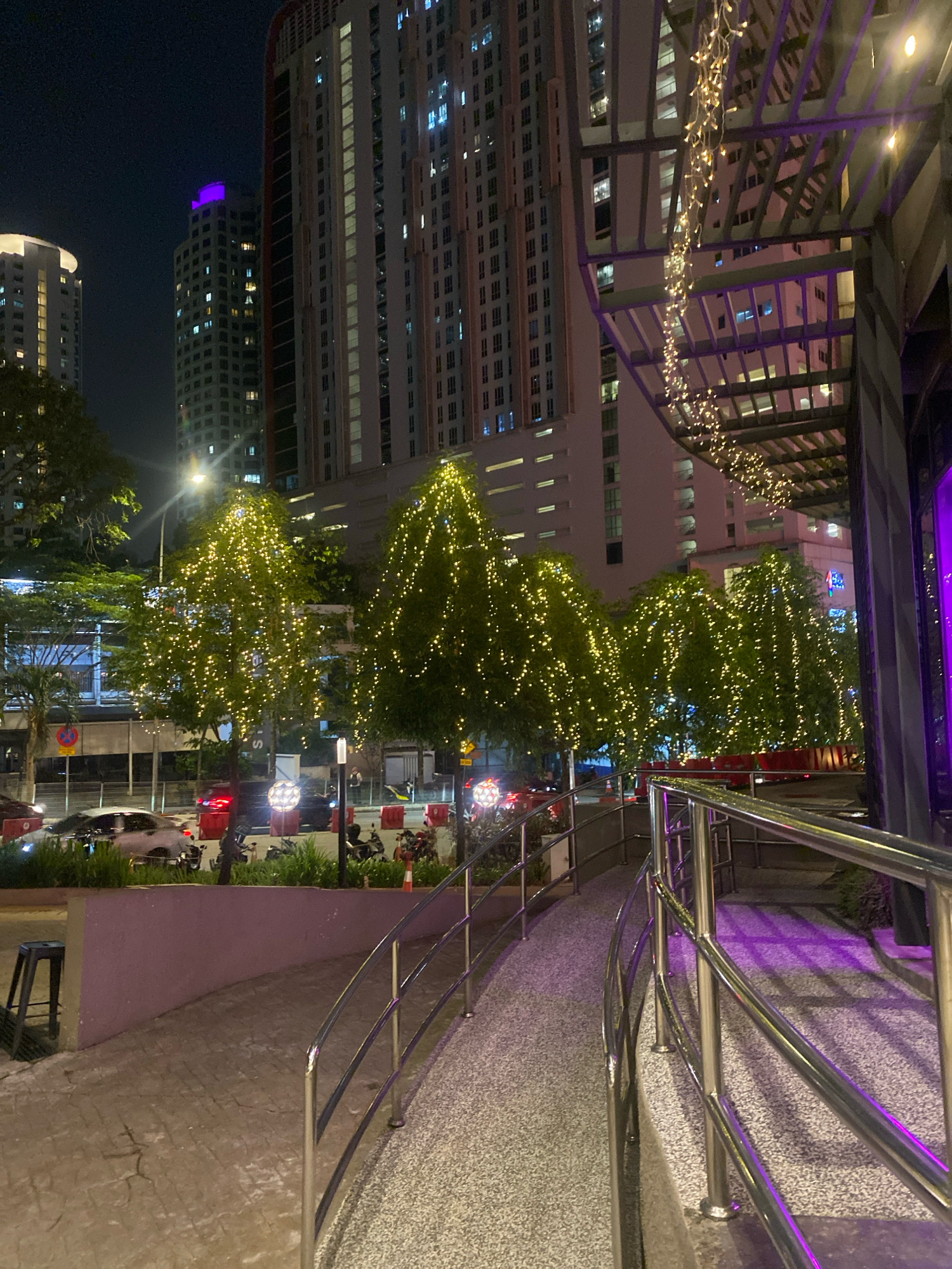 The image depicts a nighttime scene in Kuala Lumpur, Malaysia, focusing on a pedestrian walkway with decorative lights. Tall, modern buildings with numerous illuminated windows dominate the background, creating a cityscape at night. In the foreground, trees are adorned with strings of bright yellow lights, casting a festive glow. A metal railing lines the curved walkway, reflecting purple light from an unseen source. Cars are visible on a street to the left, with red taillights indicating traffic. The ground is a textured pavement. The overall atmosphere is urban and festive, suggesting a commercial or entertainment district.