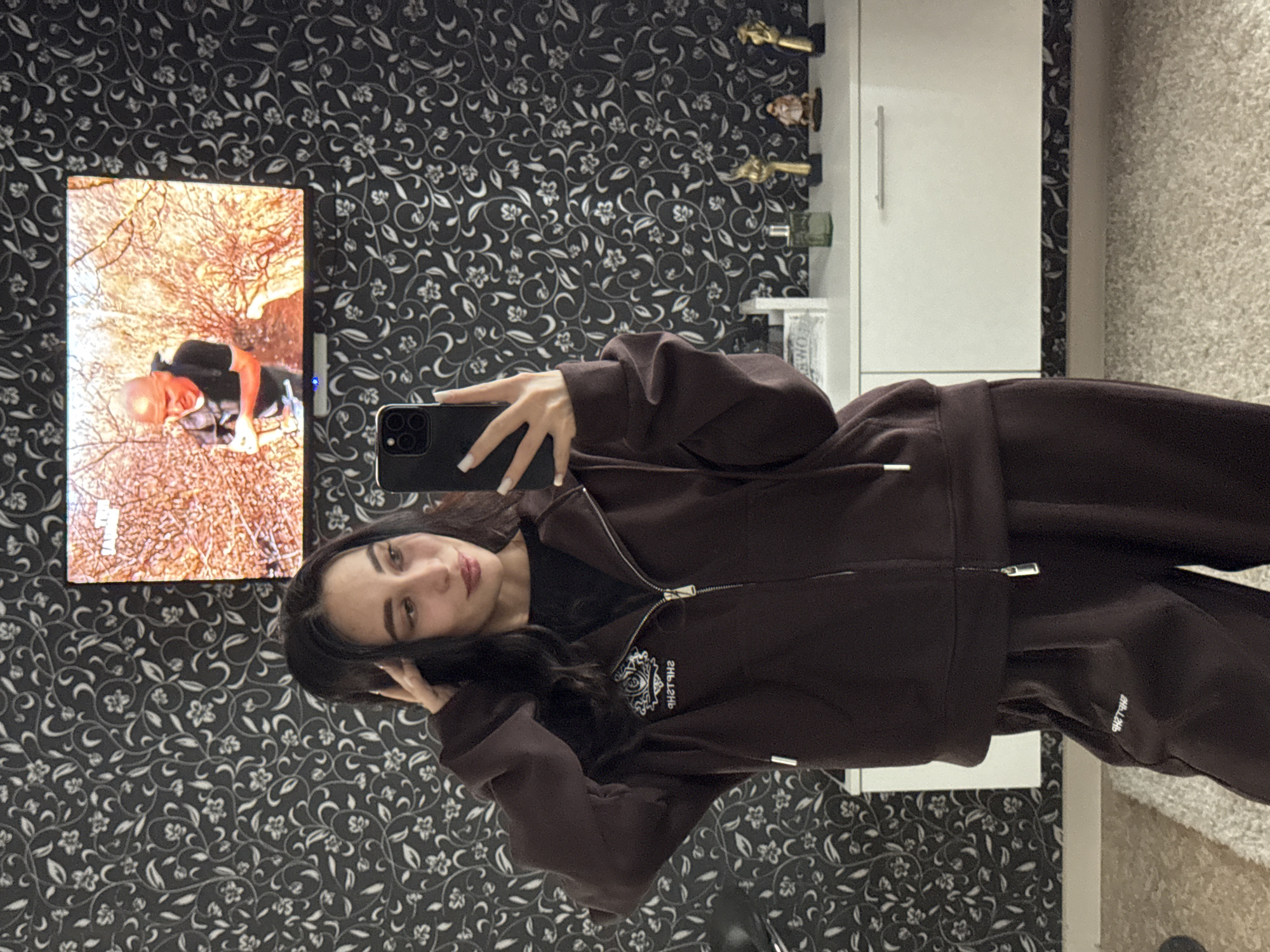 A woman is taking a selfie in what appears to be an indoor environment. She is holding a black smartphone in her right hand, and her left hand is touching her hair. She is wearing a dark brown zip-up hoodie with some sort of logo on it and matching pants. The background consists of a wall with a floral wallpaper pattern, a TV screen displaying a scene of someone walking through trees, and a white cabinet. The floor seems to be covered with a light gray rug. There is no definitive information available to confirm the location in the city of Chaikovsky, Russia, based on the provided image.