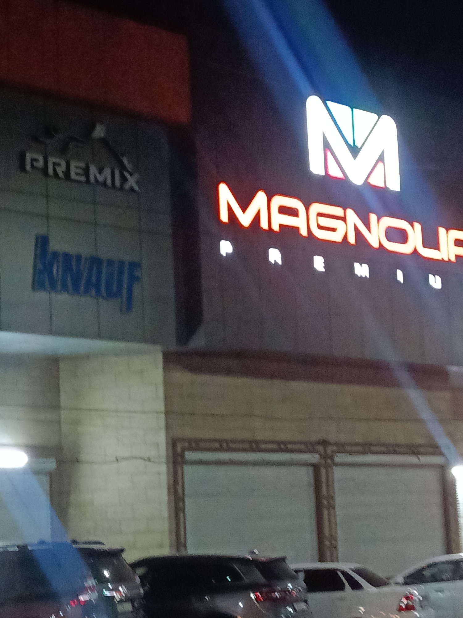 The image shows a building at night, with illuminated signage. The building features distinct signs for "PREMIX" and "KNAUF" on the left side, and a large, illuminated sign for "MAGNOLIA PREMIUM" on the right. The "MAGNOLIA PREMIUM" sign includes a prominent "M" logo. In front of the building, several vehicles are parked. The overall environment suggests a commercial or industrial area. Based on the presence of these brands, the location is identified as a Magnolia store in Shymkent, Kazakhstan.