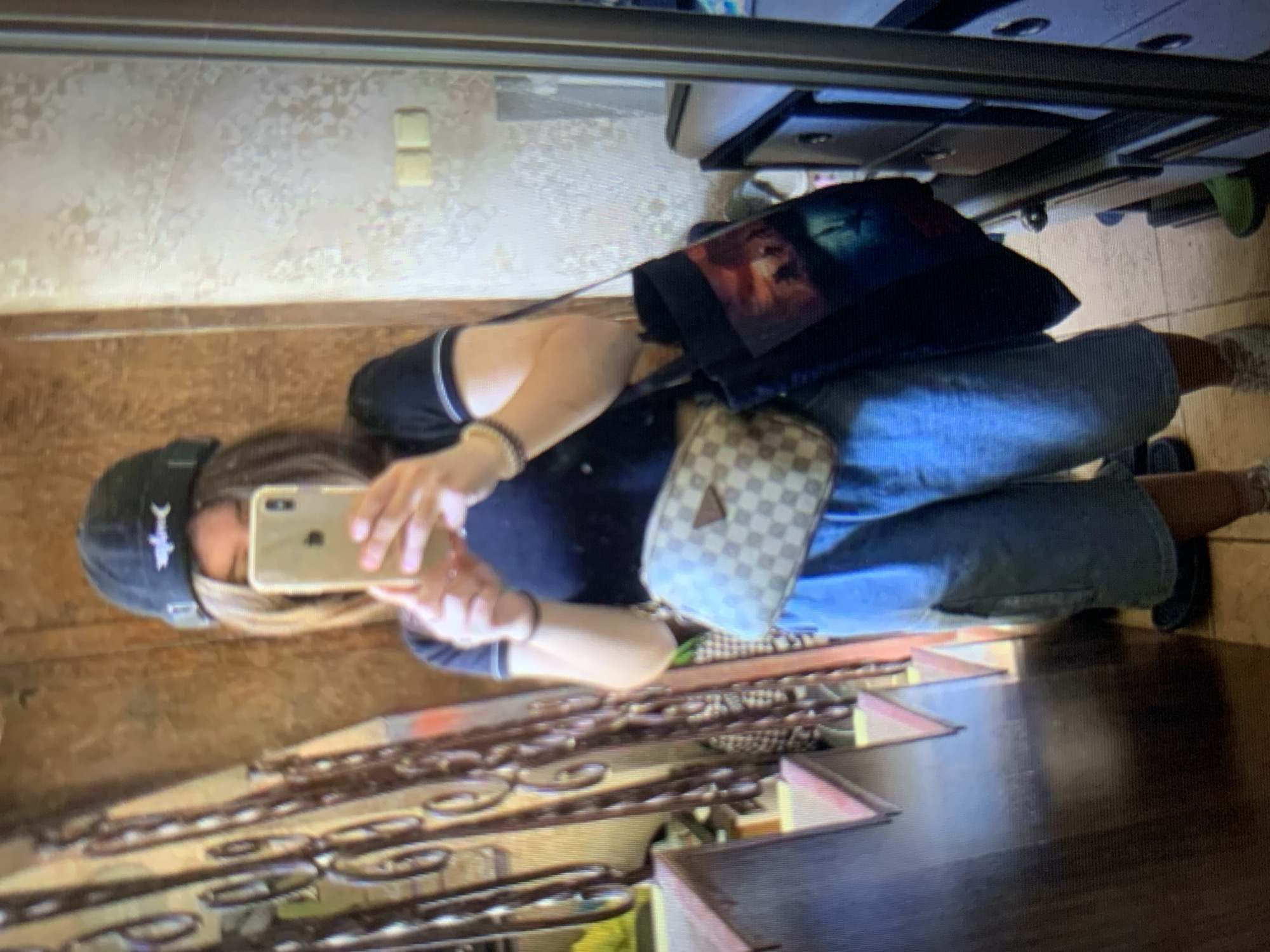 The image shows a person taking a mirror selfie indoors. The person is wearing a black baseball cap, a black t-shirt, denim shorts, a checkered fanny pack, and white sneakers. They are holding a smartphone in their right hand, capturing their reflection. A dark tote bag with a graphic print is slung over their left shoulder. The background reveals a staircase with metal railings, textured wallpaper, and a dresser or cabinet. The setting appears to be a residential interior, potentially in Lymanka, Ukraine, given the context provided. There is no visible indication of the city's specific location within the image itself.