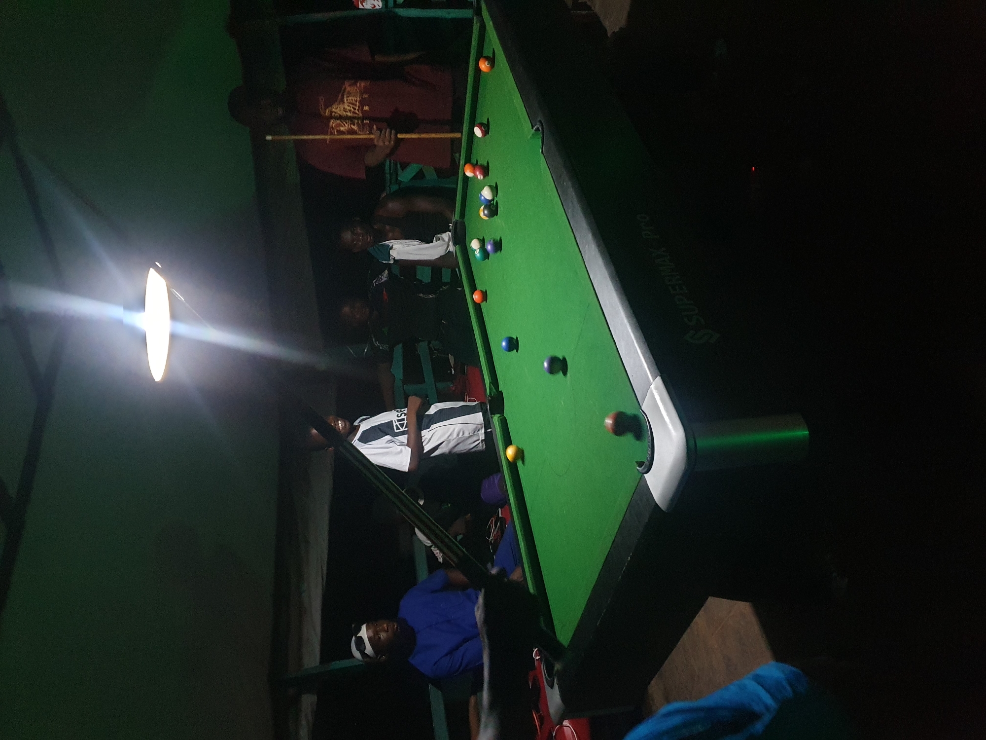The image depicts a group of young men gathered around a green pool table in an outdoor, dimly lit setting. A single overhead lamp illuminates the table and some of the individuals. Several pool balls of various colors are scattered on the table, some in play and others seemingly waiting to be struck. The pool table itself is branded with "SUPERMAX Pro". One individual on the right is holding a pool cue, suggesting a game is in progress. The environment appears to be a casual gathering place, possibly a backyard or a local hangout, typical of informal social spaces in Lokoja, Nigeria. The individuals are casually dressed, and the overall atmosphere suggests camaraderie and leisure.