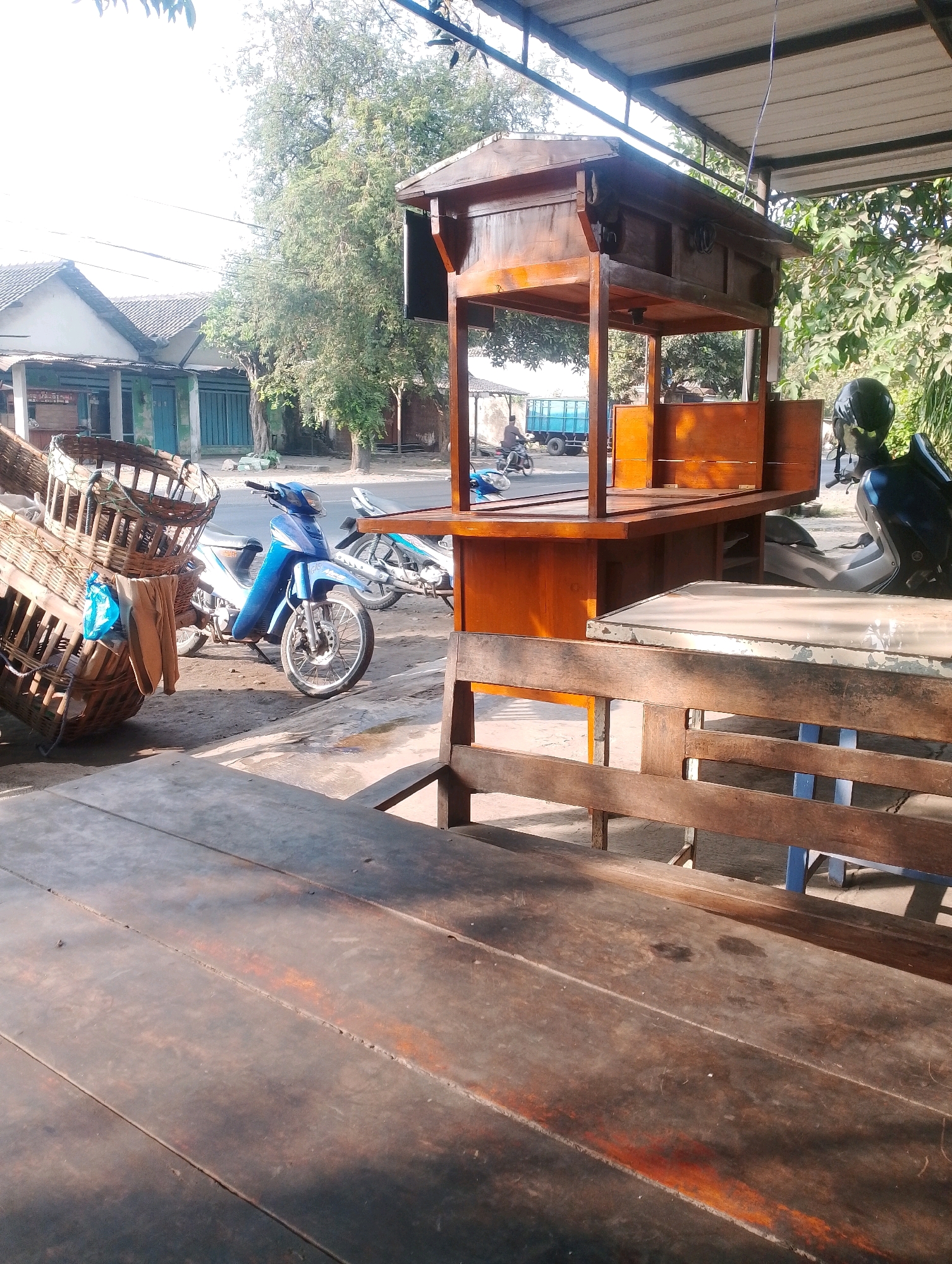 The image depicts a street scene in Jambean, Indonesia. In the foreground, a rustic wooden table and bench are visible. Behind the seating, a wooden food stall with a peaked roof stands open, displaying shelves within. To the left of the stall, two large wicker baskets are stacked precariously. A blue motorcycle is parked in front of the stall, with another motorcycle further in the background. The backdrop reveals a row of simple buildings, some with green shutters, and trees lining the street. A person is seen riding a motorcycle in the distance. The environment appears to be an outdoor, possibly commercial area on a sunny day.