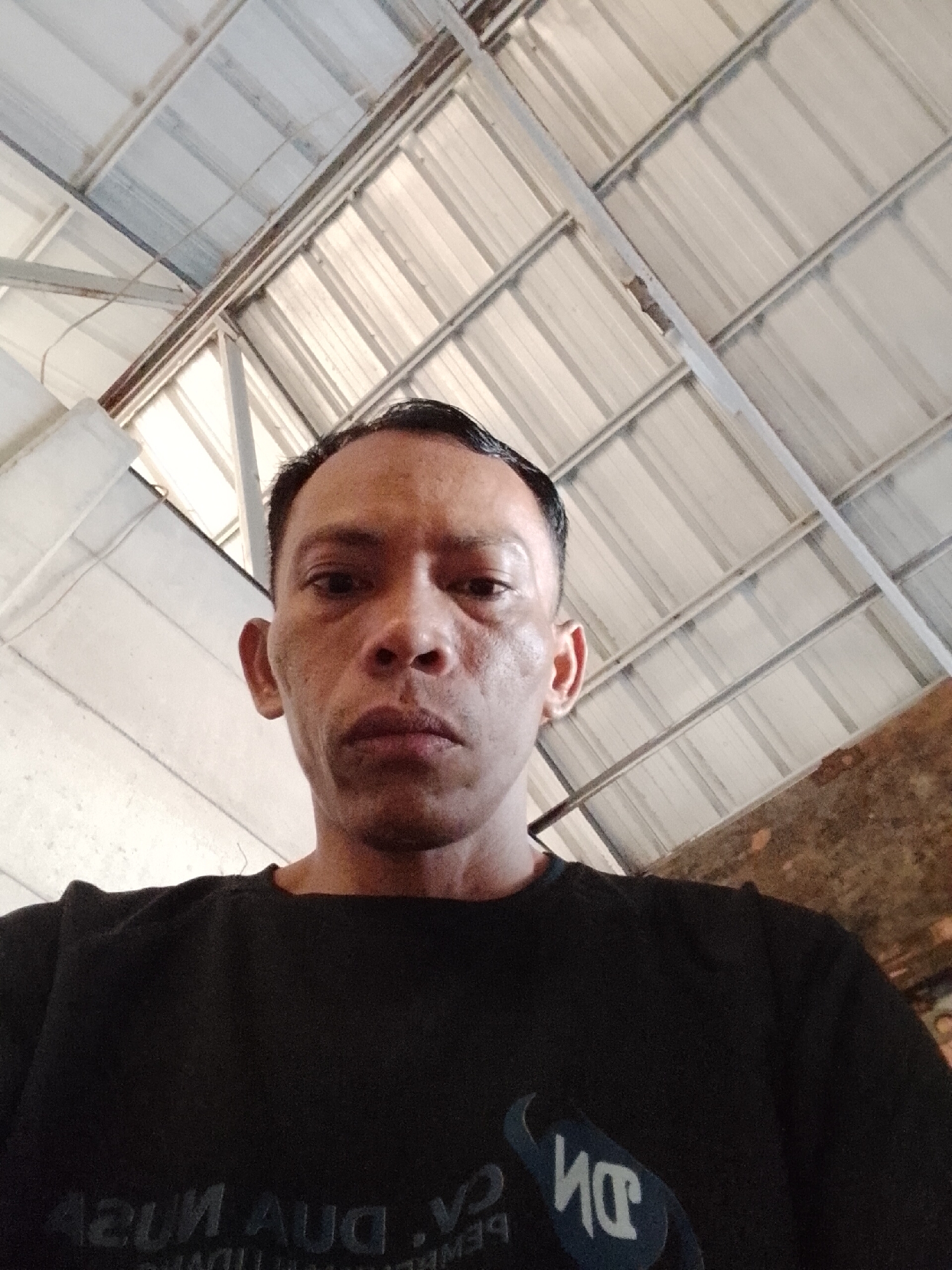 A man in a black t-shirt is looking up at the camera, with the corrugated metal roof of a building overhead. The man's face is in focus and well-lit, showing his skin texture and features. His hair is short and dark. The background consists of the metallic roof structure and some white walls. The setting appears to be an outdoor or semi-outdoor space with exposed roofing. The man is positioned in the center of the frame, taking up a significant portion of the image.