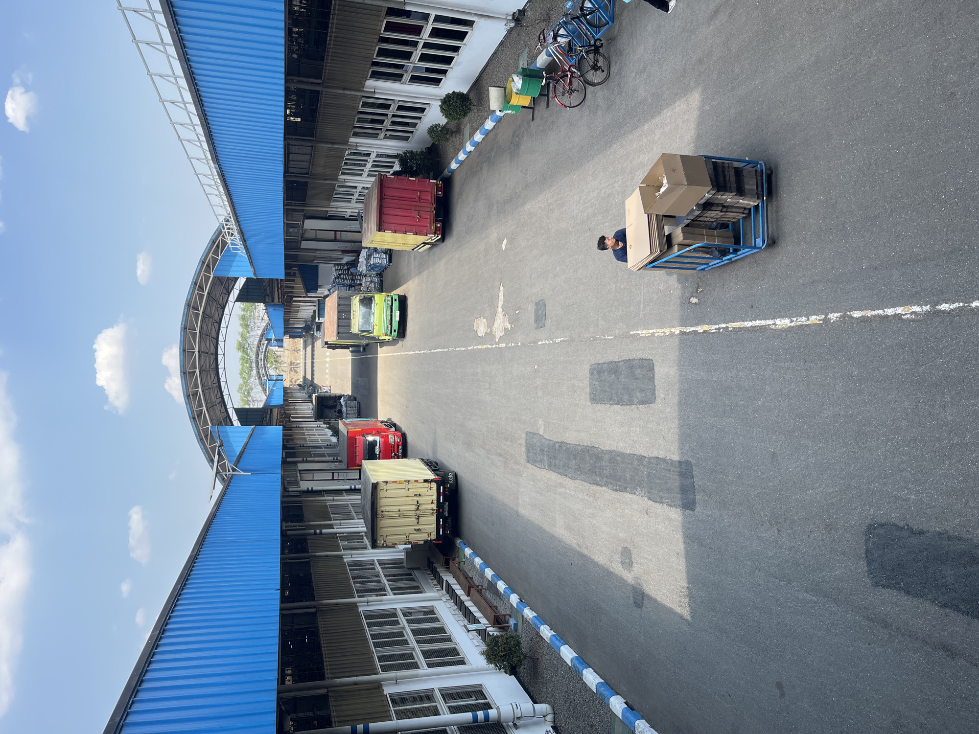 The image depicts an outdoor industrial setting with a wide asphalt road leading between two long, blue-roofed buildings. The buildings appear to be warehouses or manufacturing facilities, with rows of windows and loading bay doors visible. Several trucks are parked along the road, some loaded with containers and others appearing to be delivery trucks. In the foreground, a person is pushing a blue cart filled with cardboard boxes. The sky is blue with scattered white clouds, indicating a daytime scene. The environment appears to be clean and orderly, suggesting an operational industrial zone. The location is identified as Pelang, Indonesia, though no specific landmarks within the city are visible.