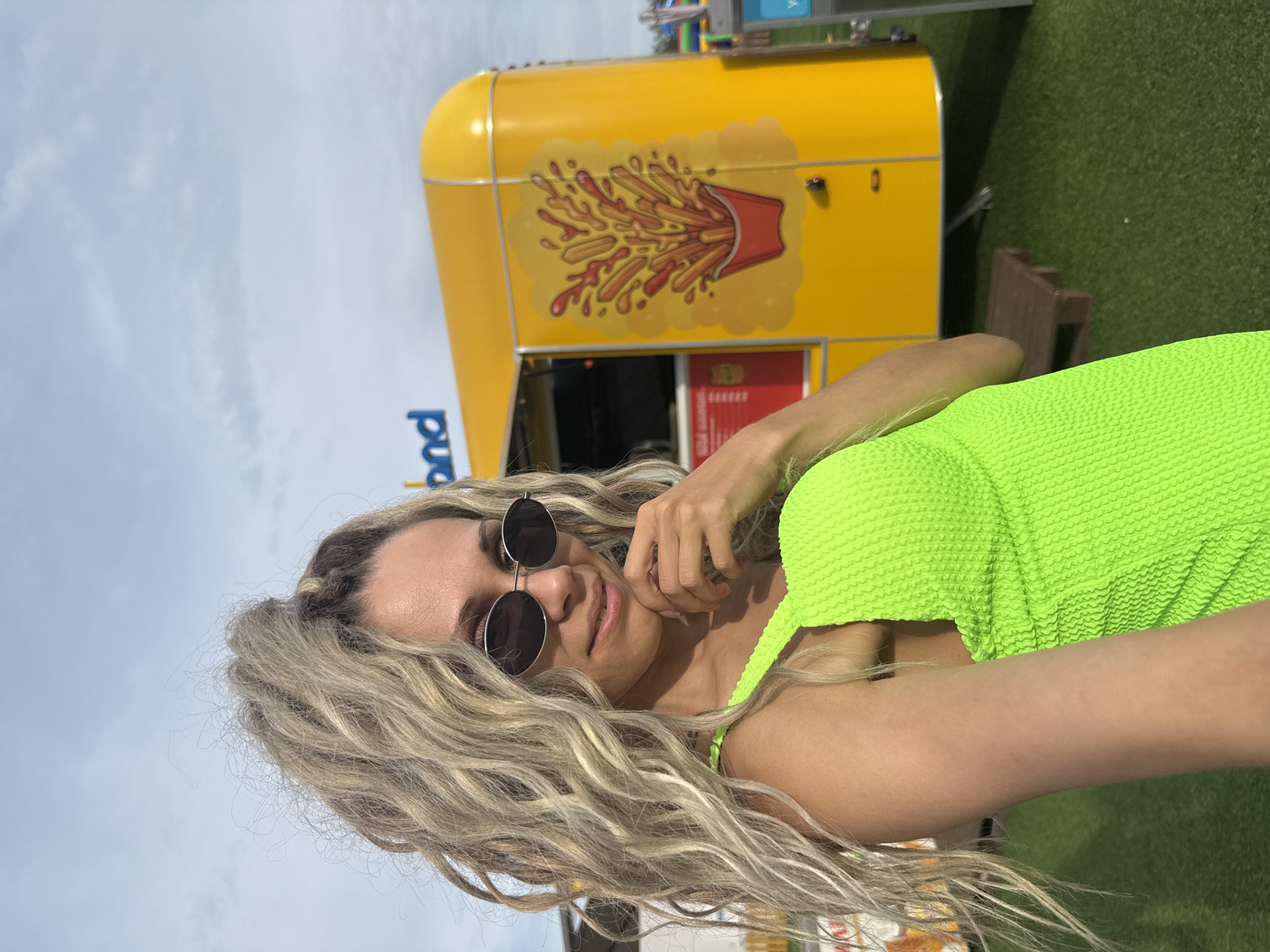 A woman with blonde, curly hair tied up in a ponytail and wearing oval sunglasses and a neon green swimsuit poses for a selfie. She is outdoors, with a bright yellow food truck behind her. The food truck has a cartoon illustration of french fries with ketchup. The ground is covered in green artificial turf. The sky is overcast. A sign is visible on the food truck that says "band".