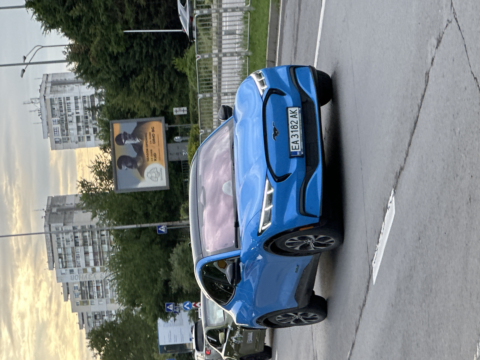 The image captures an urban street scene in Varna, Bulgaria, featuring a vibrant blue Ford Mustang Mach-E electric SUV. The car is prominently displayed on the road, with its front license plate "BG EA 3182 AK" clearly visible. The time of day appears to be late afternoon or early evening, suggested by the diffused light and the warm, lighter hues of the sky reflecting on distant surfaces.
In the background, tall, concrete apartment buildings, characteristic of post-socialist architecture, line the street, partially obscured by green trees. One building distinctly features the Cyrillic text "ДОНАЕВ" (Donayev) painted vertically on its side. Streetlights, metal barriers, and various traffic signs are also present along the roadside.
A large billboard stands in the mid-ground, displaying an advertisement with two figures wearing hats and suits. Visible text on the billboard includes "SEASON OPENING", "23.05.2018", "DINO BG", and "KAD", accompanied by a circular logo. The road surface, made of asphalt, shows some cracks and white lane markings. Other vehicles can be faintly seen further down the road. The scene portrays a bustling urban environment.