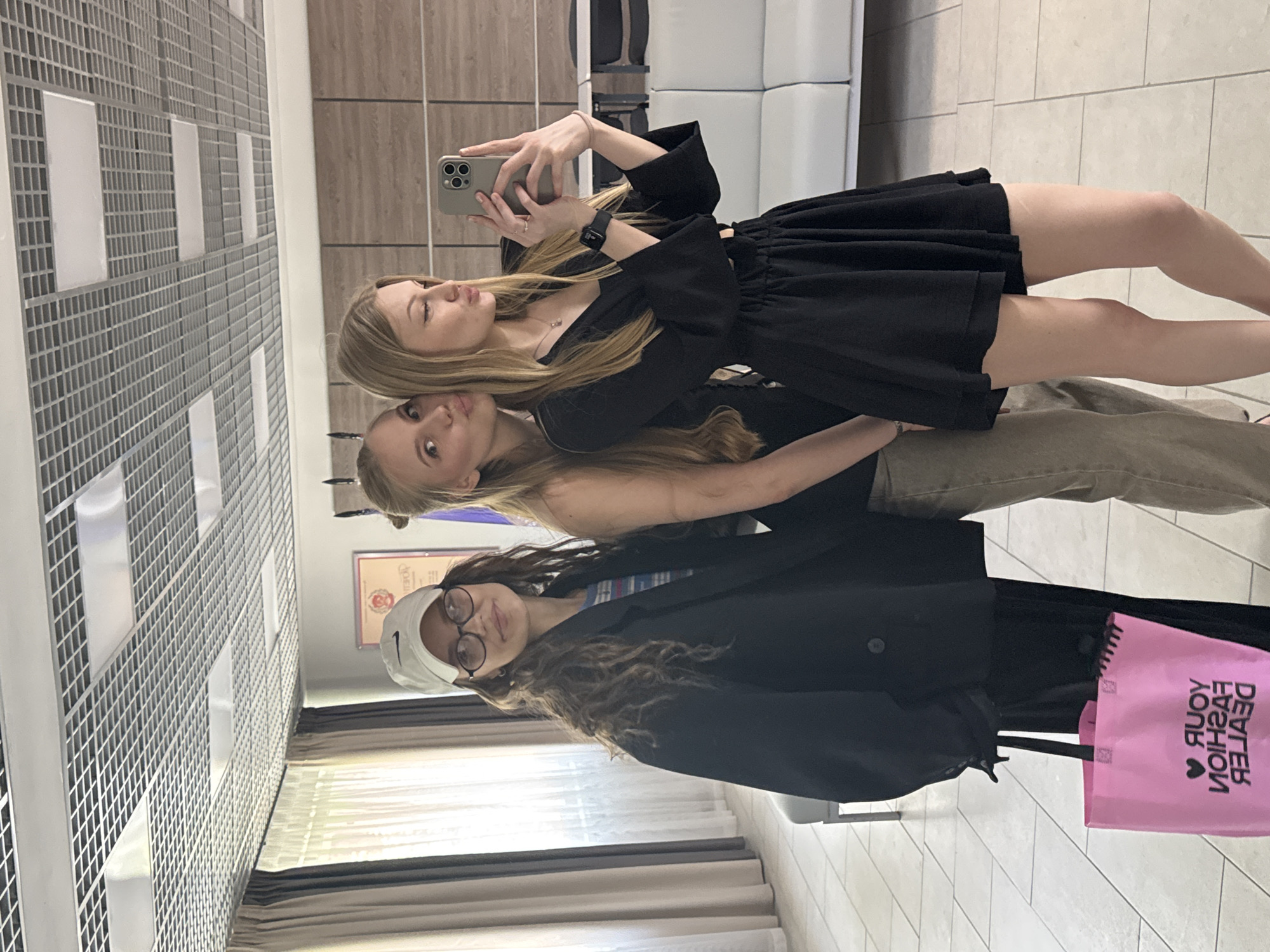 Three women are standing together in what appears to be an indoor space, possibly a retail setting, taking a selfie in a mirror. The woman on the top is holding a smartphone and seems to be taking the picture. She is wearing a black dress. The woman in the middle has long blonde hair and is wearing pants. The woman at the bottom is wearing a white baseball cap, glasses, and a black jacket. She has a pink shopping bag, which has backward text. The background shows a wall with a tiled pattern, a wooden wall, and a white sofa. The text in the image on the pink bag reads "YOUR FASHION DEALER"
The geographical location is Perm, Russia.