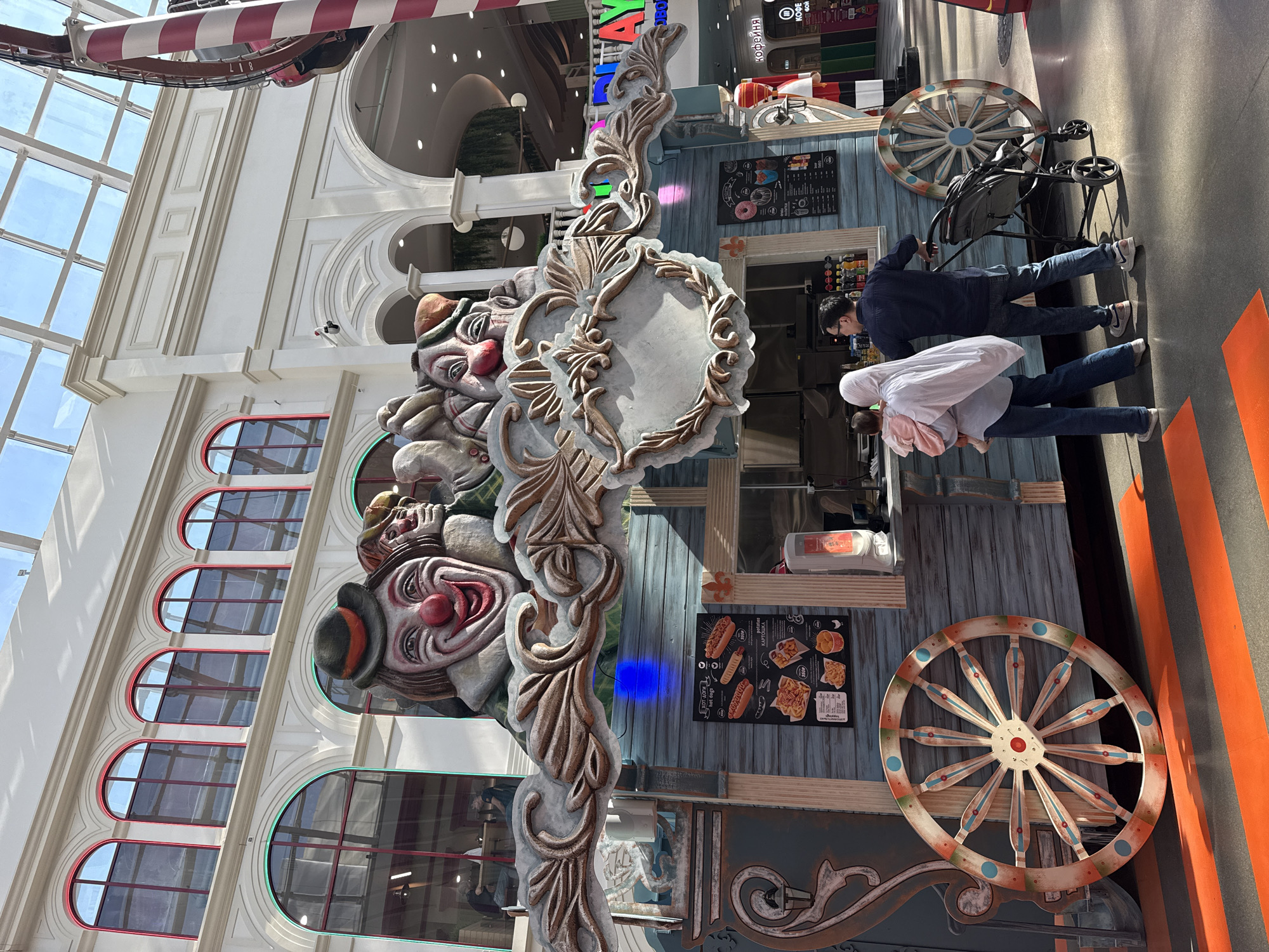 The image shows a kiosk in what appears to be a shopping mall. The kiosk is decorated with a large clown-themed facade. Two people are standing at the kiosk, seemingly placing an order. A stroller is parked nearby. The environment seems to be an indoor mall with various architectural details, including large windows, arched openings, and structural elements. A sign with Russian text suggests this could be in Moscow, Russia.