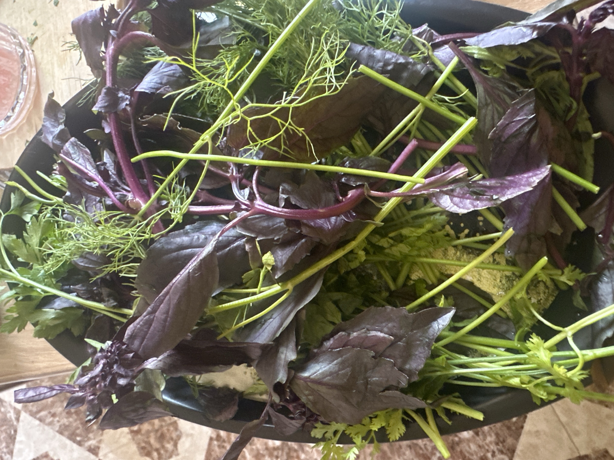 A close-up, overhead shot shows a bounty of fresh herbs in a dark bowl. The herbs include deep purple basil leaves with vibrant green stems, feathery dill fronds, and the lighter green, finely chopped leaves of cilantro. The arrangement is lush and appears to be recently harvested, with some dew or moisture visible on the leaves and stems. The background is indistinct, with a suggestion of a patterned surface and a blurred glimpse of a glass with pink liquid. The overall impression is one of freshness and natural abundance.