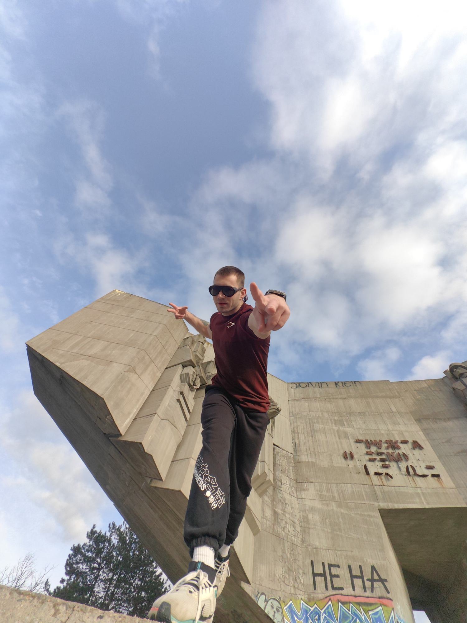 A low-angle shot captures a man in front of the Bulgarian-Soviet Friendship Monument in Varna, Bulgaria. He is wearing a burgundy t-shirt, dark sweatpants with text on the side, sunglasses, and sneakers. He is pointing directly at the camera with his right hand and holding up a peace sign with his left. He is standing on a ledge, with the monument's concrete structure behind him. Graffiti is visible on the monument, and the sky is cloudy. A tree is visible in the lower left corner.