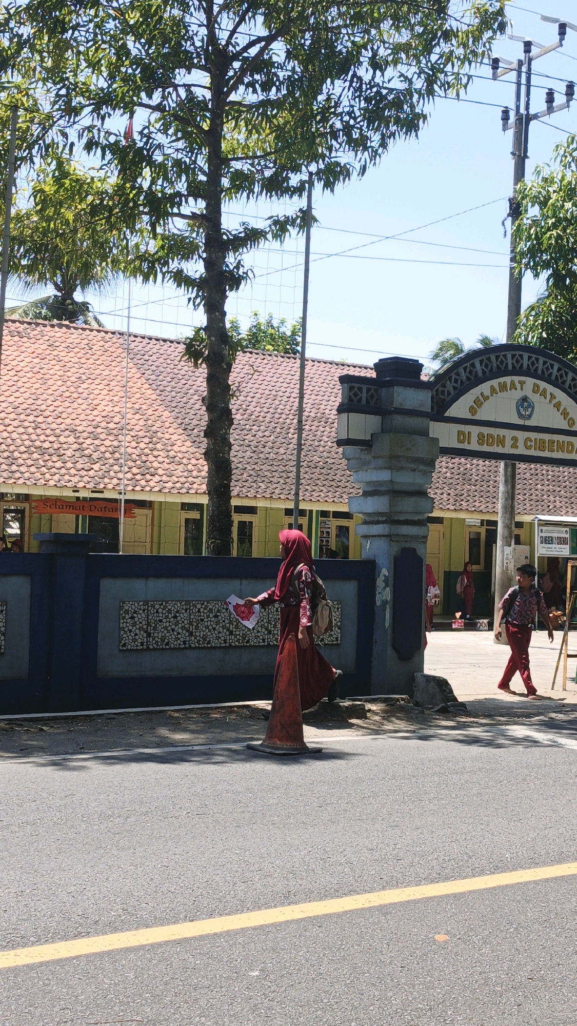 The image shows the entrance to an educational institution in Cibenda, Indonesia. A young person wearing a red hijab and a red dress walks towards the right side of the frame, holding a white and red paper. Behind them, a young boy in a red school uniform also walks towards the right. The school building has a red-tiled roof and yellow walls. The entrance features a decorative archway with the text "SELAMAT DATANG DI SDN 2 CIBENDA" (Welcome to Cibenda Public Elementary School 2). The area appears to be a street with asphalt pavement and a yellow dividing line. A traffic cone is visible on the side of the road in front of the school. Trees and utility poles are present in the background, indicating an outdoor, daytime scene under a clear sky.