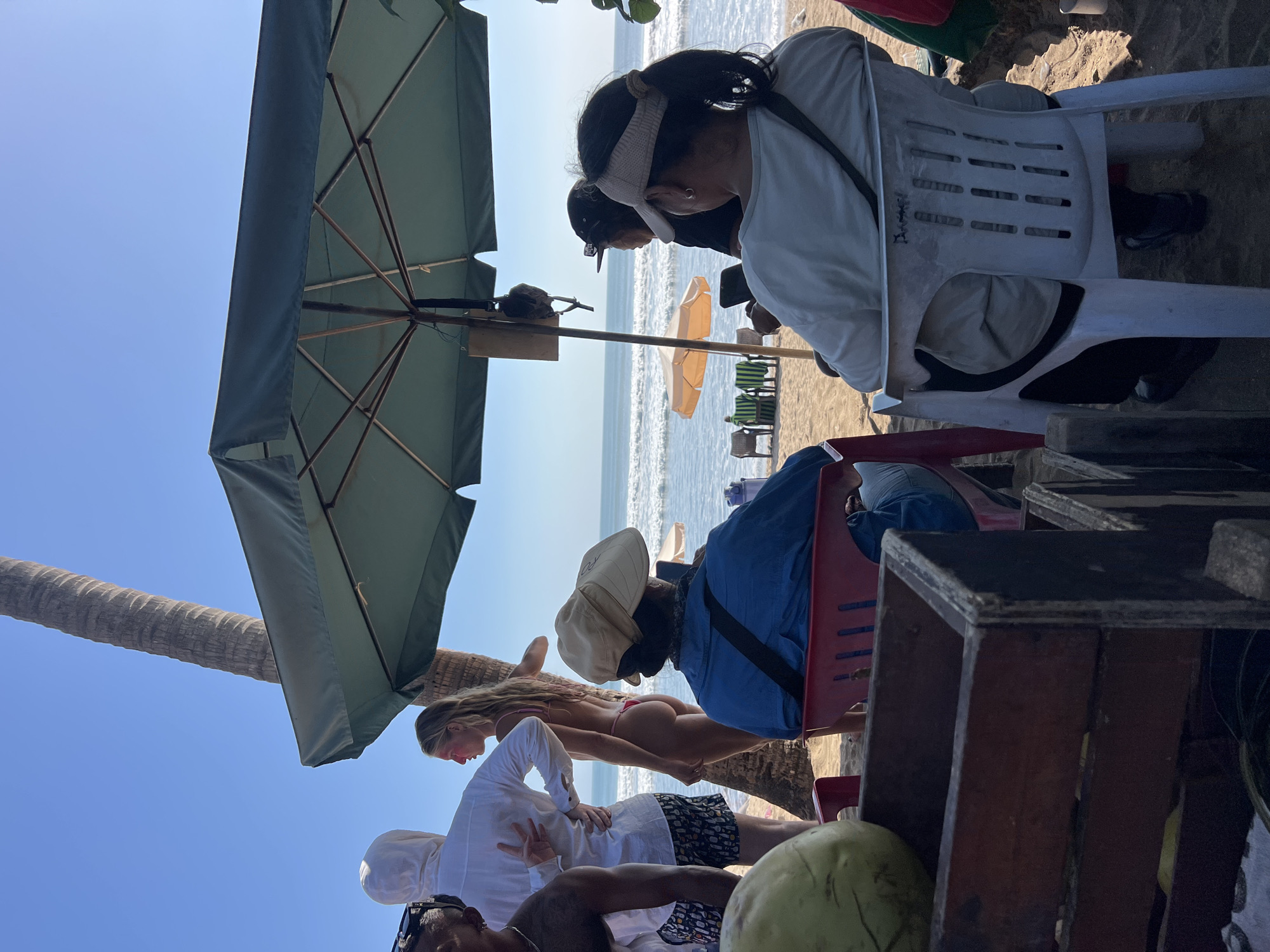 In Kuta, Indonesia, on a sunny day at the beach, a group of people are gathered under a large green umbrella. In the foreground, a person wearing a blue jacket and a beige hat sits in a red plastic chair, facing away from the camera, appearing to look at a phone. To their right, another person, wearing a light-colored jacket and a visor, also sits in a white plastic chair, looking out towards the ocean.
Behind them, a man in a white hoodie and patterned shorts stands with his arm around a woman wearing a pink bikini. The woman has her back to the camera, her hair long and blonde, and she is looking out at the sea. Another person, partially visible, stands to the left of the man in the white hoodie.
In the mid-ground, several empty chairs and a small orange umbrella are set up on the sand, with the ocean waves gently rolling in. A tall palm tree stands to the right of the main group, its trunk prominent against the clear blue sky. In the far left foreground, a portion of a wooden crate containing a large green coconut is visible. The sand is a light brown, and the overall atmosphere suggests a relaxed beach scene.