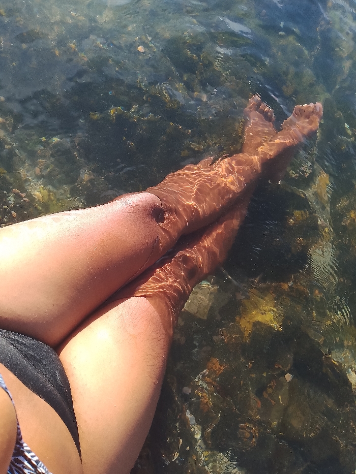 A sunlit shot shows legs submerged in clear, shallow water. The legs are tanned and partially submerged, with feet crossed near the surface. The water distorts the limbs, creating ripples and reflections. Below, the seabed is visible with rocks and pebbles. A portion of swimwear is visible in the lower-left corner.