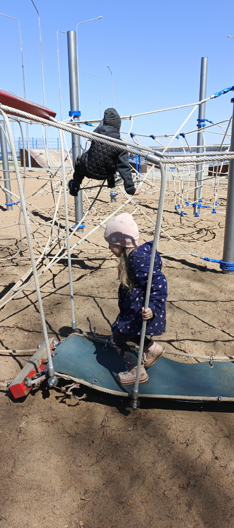 Two children play on a playground obstacle course in Chaikovsky, Russia. The playground has metal poles, sand, and a net of ropes. One child wearing a dark coat and hat climbs upside down on the rope net, while the other child wearing a patterned coat and a pink hat stands on a blue suspended platform. The child on the platform holds onto the rope supports.