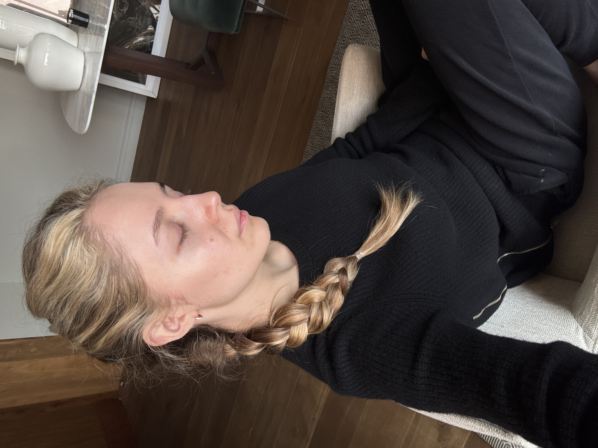 A woman with fair skin, light brown hair braided, and wearing a black sweater and black pants is lying on a beige chair. The setting appears to be indoors. The scene includes a white table with a white vase on top, a dark green chair, and wooden floors. There is a picture frame on the table. The image does not provide information to ascertain the location is Mexico City, Mexico.