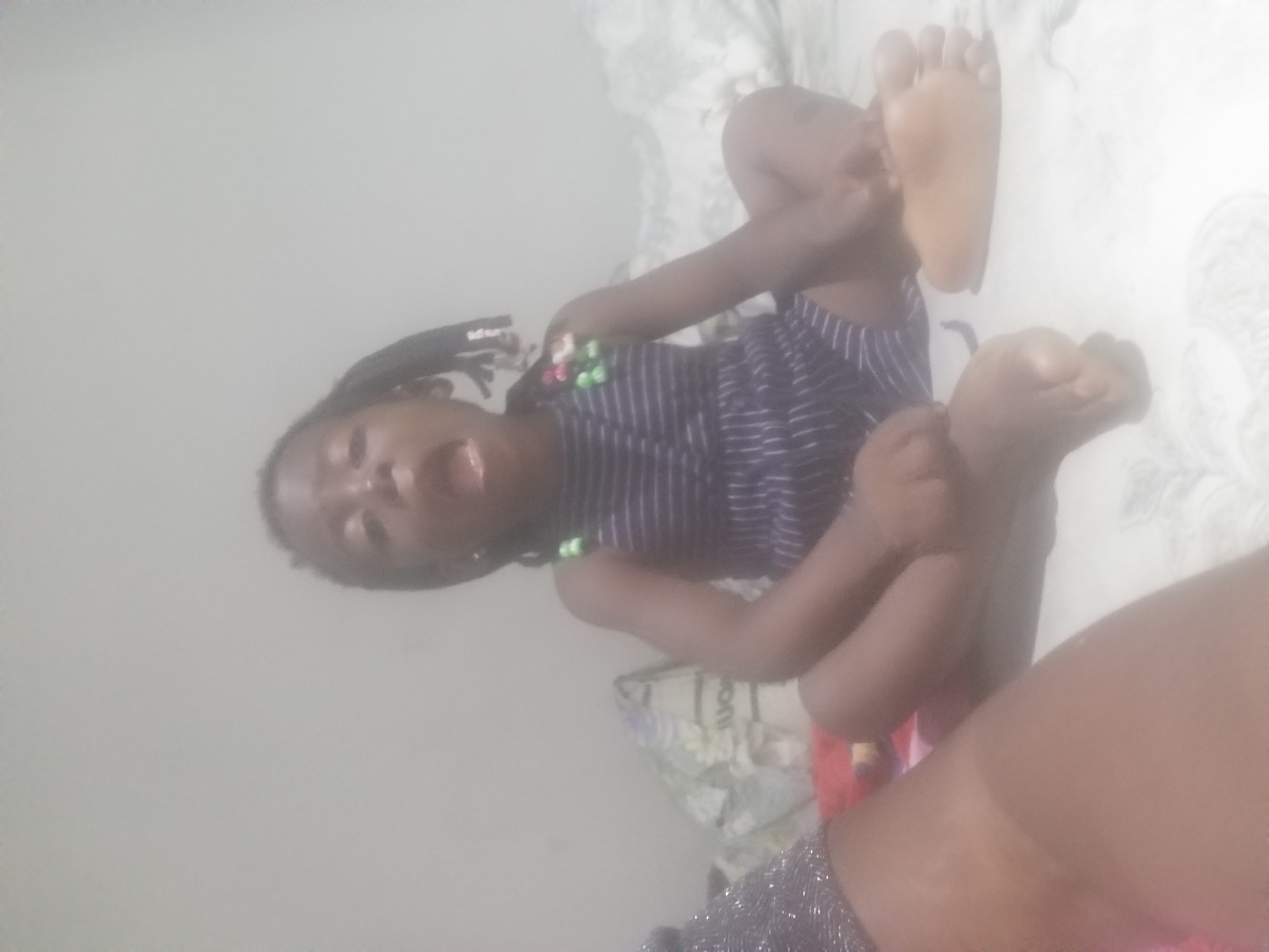 The image shows a young Black child sitting on a bed with their mouth open, appearing to be speaking or shouting. The child is wearing a dark blue and white striped dress. The background is a plain, light-colored wall. The overall scene is blurry and lacks clear details about the environment, making it impossible to determine the location, including whether it is in Kano, Nigeria.
