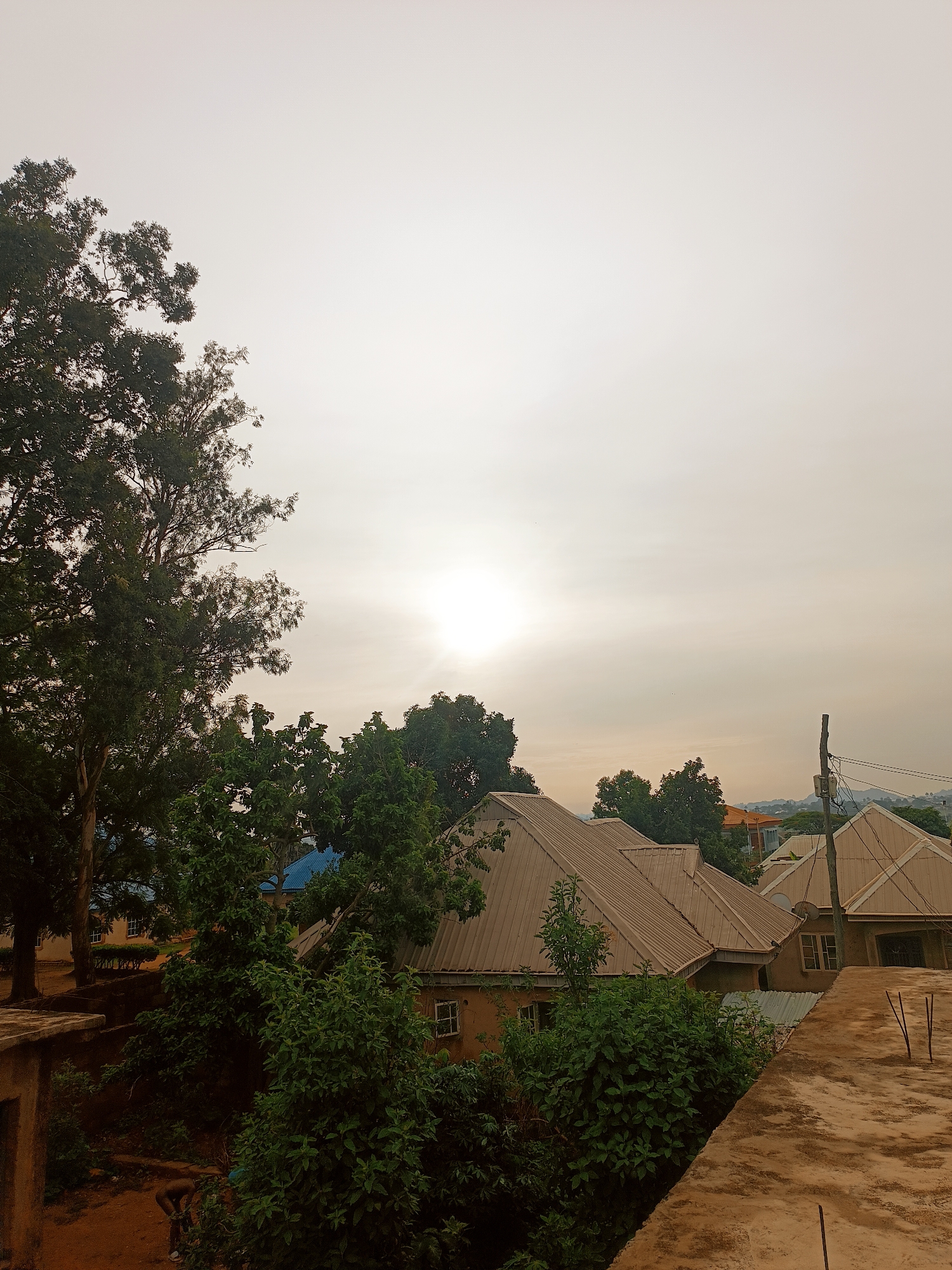 The image depicts a residential area in Bukuru, Nigeria, taken from an elevated perspective, possibly a rooftop. The foreground features a weathered concrete surface. Lush greenery, including trees and bushes, fills the middle ground. Several single-story buildings with metal roofs are interspersed among the foliage. In the background, a hazy sky obscures the sun, creating a diffused light. Utility poles and distant hills are faintly visible. The scene suggests a quiet, suburban environment on an overcast day. There is no evidence of people in the image.