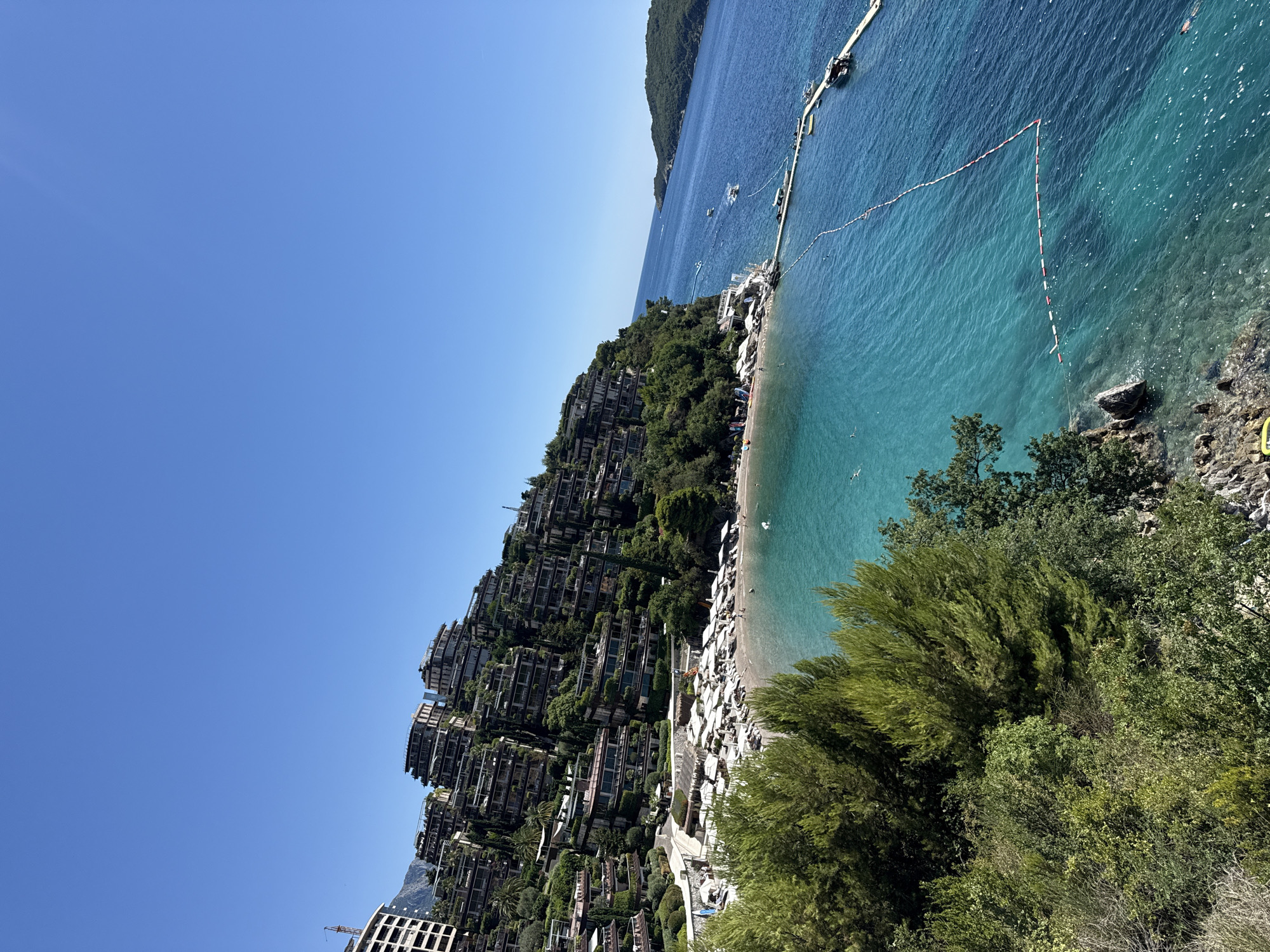 This aerial, eye-level, wide-angle shot captures a sun-drenched resort complex built into a hillside overlooking a clear turquoise bay. The resort's architecture features multiple terraced levels of buildings, each with numerous balconies, designed to blend with the lush green vegetation that covers the slope. Below the resort, a narrow, pebbly beach is lined with rows of white umbrellas and sun loungers, with a few scattered beachgoers visible. The bay itself is a vibrant blue-green, with calm water and a floating dock extending into the sea. A few small boats are moored near the dock, and a rope barrier with buoys demarcates a swimming area. In the distance, a mountainous island covered in green foliage forms the horizon. The sky is a clear, cloudless blue, indicating fair weather. The scene suggests a luxurious coastal vacation destination. The location is likely the Dukley Gardens resort in Budva, Montenegro, given its characteristic terraced buildings cascading down to the sea.