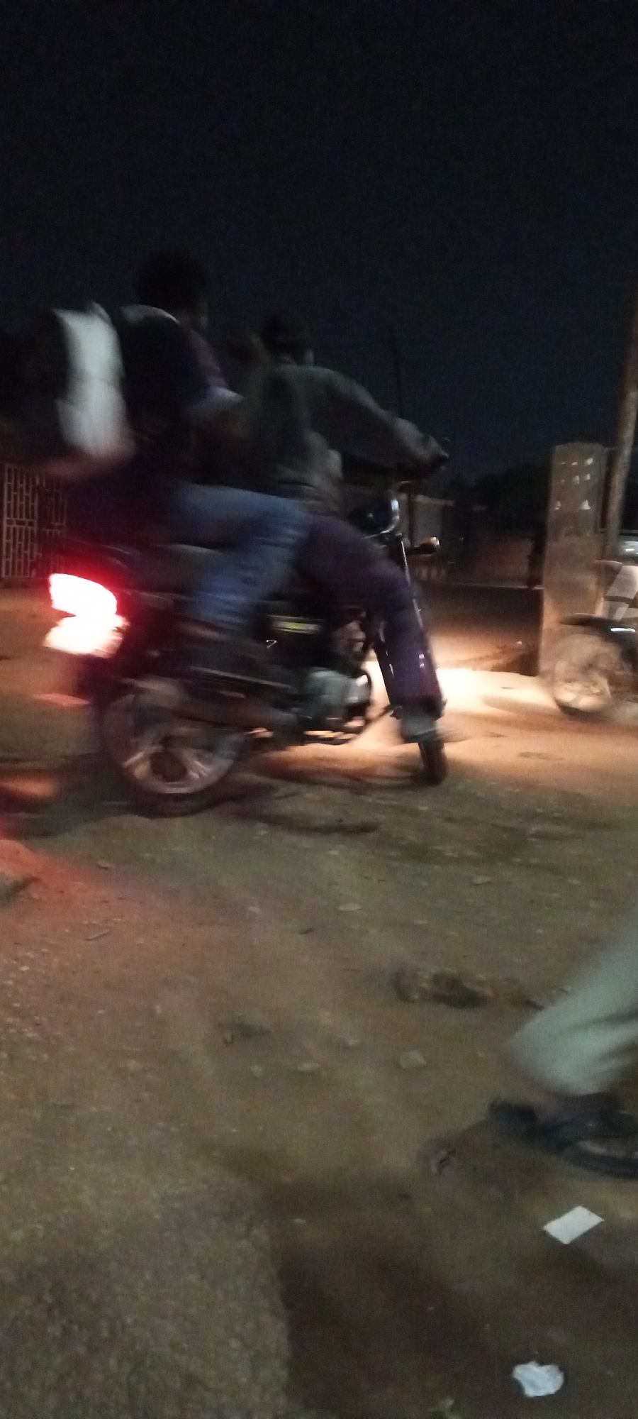 A blurred image captures two individuals riding a motorcycle at night in Sabon Tasha, Nigeria. The motorcycle's tail light is illuminated, and one rider carries a light-colored bag. Another motorcycle is parked near a structure in the background. Debris is scattered on the ground. The image is taken from a low angle.