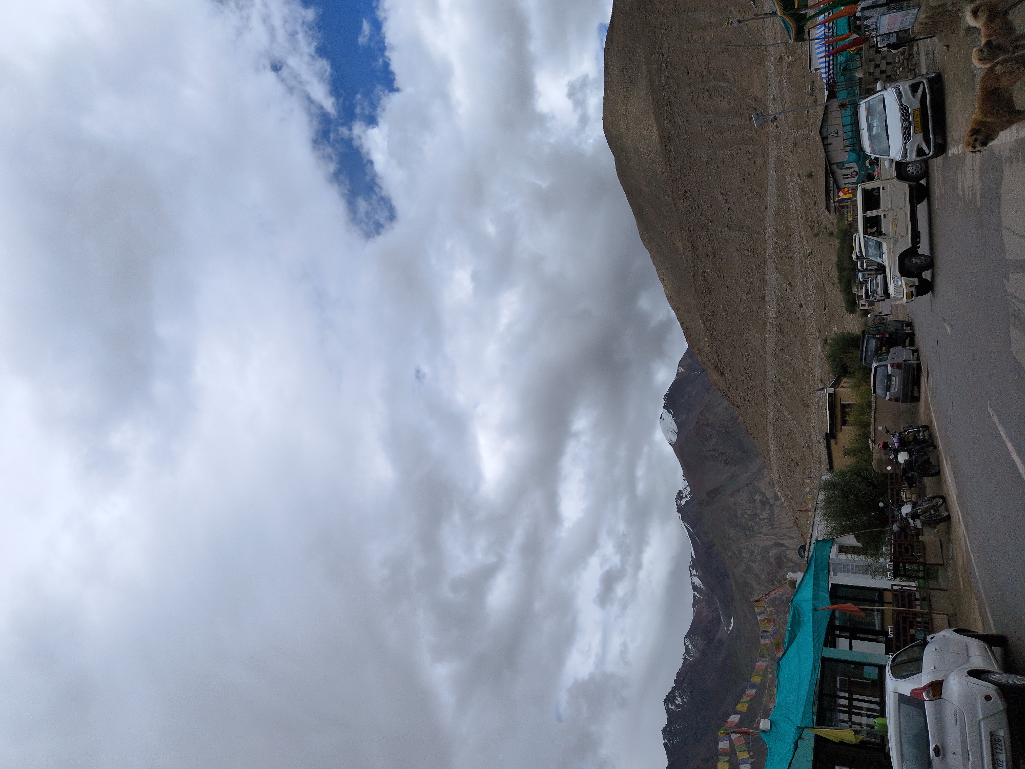 A street scene in Khardung, India, features a road with several vehicles, including a white SUV, a white utility vehicle, motorcycles, and a white car. Mountains with snow-capped peaks form the backdrop, under a cloudy sky with patches of blue. Buildings line the side of the road, some appearing to be shops or restaurants with colorful awnings. Prayer flags are strung across some of the buildings. Two dogs are visible in the foreground on the right side of the image. The overall impression is of a roadside settlement in a mountainous region.