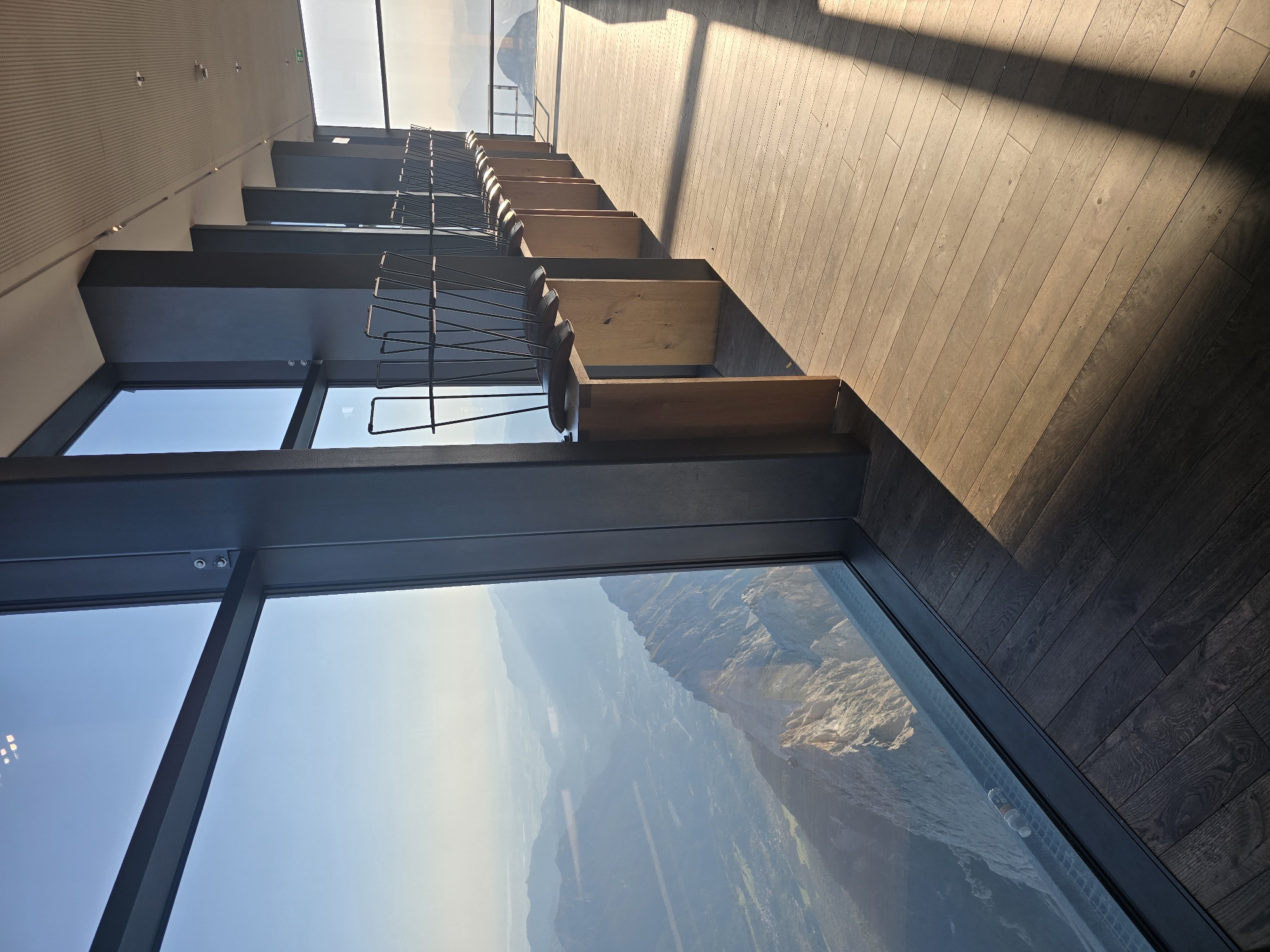 The image depicts the interior of a modern observation deck with panoramic views of the Bavarian Alps near Garmisch-Partenkirchen, Germany. A large floor-to-ceiling window dominates the left side of the frame, offering a breathtaking vista of rugged mountains and a hazy valley below. Sunlight streams into the room, casting long shadows across the polished wooden floor.
Along the right side of the room, a series of minimalist wooden stands are arranged, each topped with a dark metal chair. These chairs are stacked or placed in a way that suggests they are not in use. The architectural elements of the room are sleek and contemporary, with dark grey pillars supporting the ceiling, which features a perforated acoustic paneling. A few discreet lights are visible on the ceiling.
The view outside the window reveals a dramatic mountain landscape, with sharp, rocky peaks in the foreground and a more subdued, undulating terrain in the distance. The light suggests it is either early morning or late afternoon. No people are visible in the image. A single water bottle is visible on the windowsill.