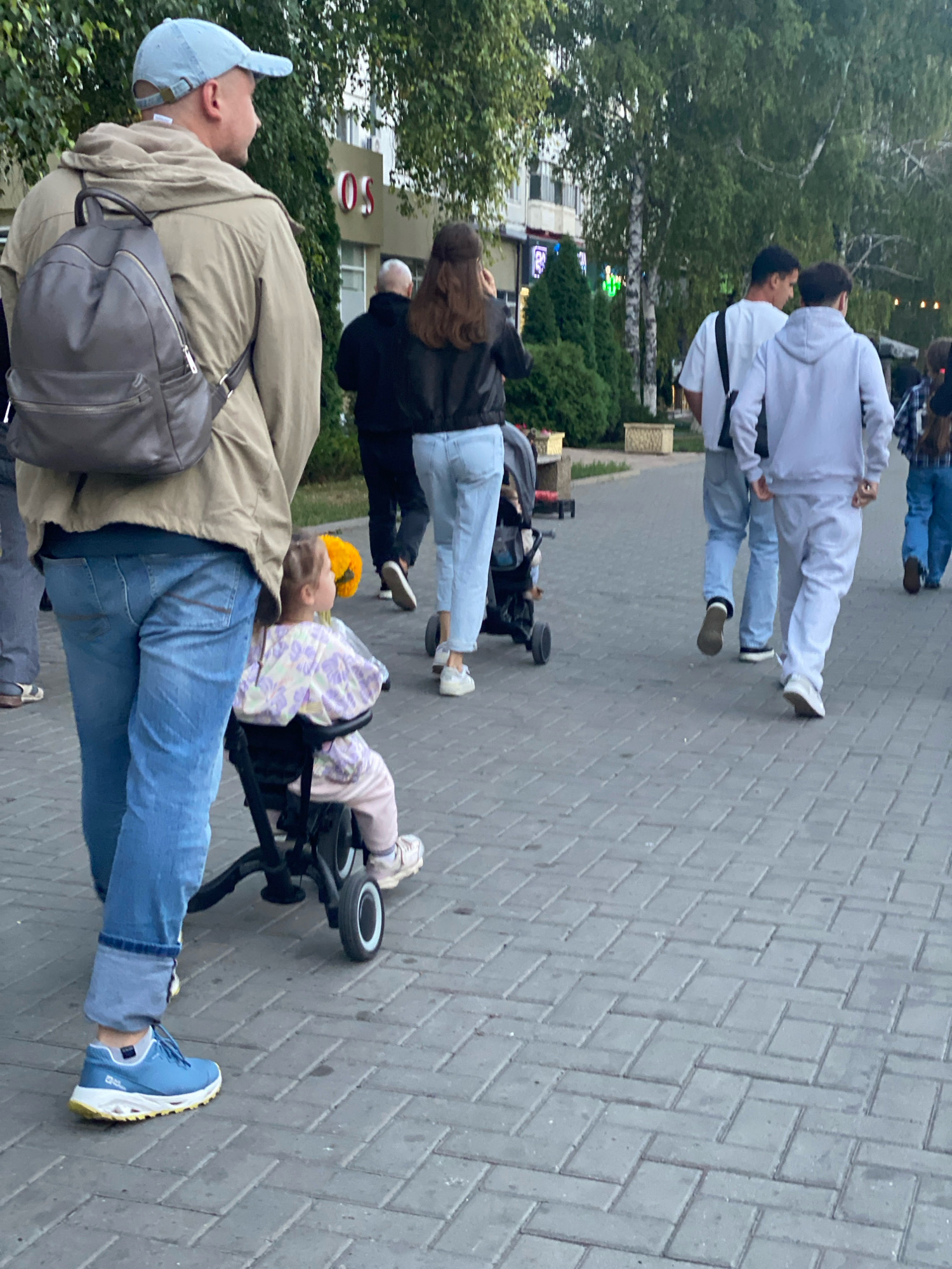 The image depicts a street scene in Chișinău, Moldova, likely in the late afternoon or early evening given the lighting. The foreground features a man from the waist down, wearing a light brown hooded jacket, blue jeans, and blue athletic shoes. He is carrying a dark grey backpack. Beside him, a young child is seated in a black tricycle, facing away from the camera. The child is wearing a light-colored, patterned top and pink pants, with a yellow object, possibly a flower, behind them.
In the mid-ground, several other individuals are walking along a paved pedestrian walkway. To the left, a person in a black top and jeans is seen from behind. Further back, two young men are walking away from the camera. One is wearing a white t-shirt and light grey pants, and the other is in a light grey hooded sweatshirt and matching pants. Another individual in a plaid shirt and jeans is also visible in the background. The walkway is made of grey interlocking bricks.
The background reveals a street lined with trees, their foliage appearing green and full. Buildings with visible signage are present, including one with red lettering that appears to spell "OS". Streetlights and other urban elements are also discernible. The overall impression is of a public urban space, likely a promenade or sidewalk in Chișinău.