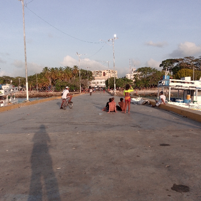 A sunlit outdoor scene shows a concrete pier with people enjoying the day. A man rides a bicycle, while a group of three people sit on the pier. A woman stands nearby in a swimsuit and crop top. A man sits on the pier's edge next to a docked boat. Streetlights line the pier. Palm trees and buildings are visible in the background under a cloudy sky. A long shadow of a person taking the photo stretches across the pier.