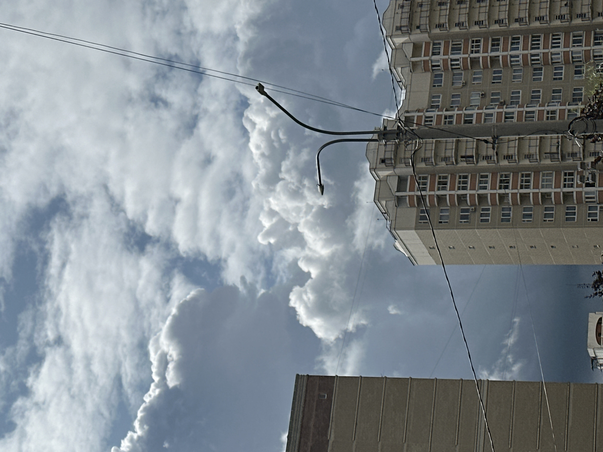 The image shows a cityscape with buildings and a cloudy sky. On the left is a portion of a beige apartment building with a dark red band near the top and small, dark rectangular windows. To the right of this building is a taller, multi-story apartment building with a light beige facade and numerous balconies with dark railings. The windows on this building are also dark and appear regularly spaced. A concrete lamppost with two curved arms supporting streetlights stands in front of the taller building, draped with electrical wires. The sky is filled with dramatic, dark gray and white cumulus clouds, suggesting potentially stormy weather. The scene appears to be an exterior view of an urban residential area, possibly in Podolsk, Russia, given the architectural style and the presence of apartment complexes. There are no people visible in the image.