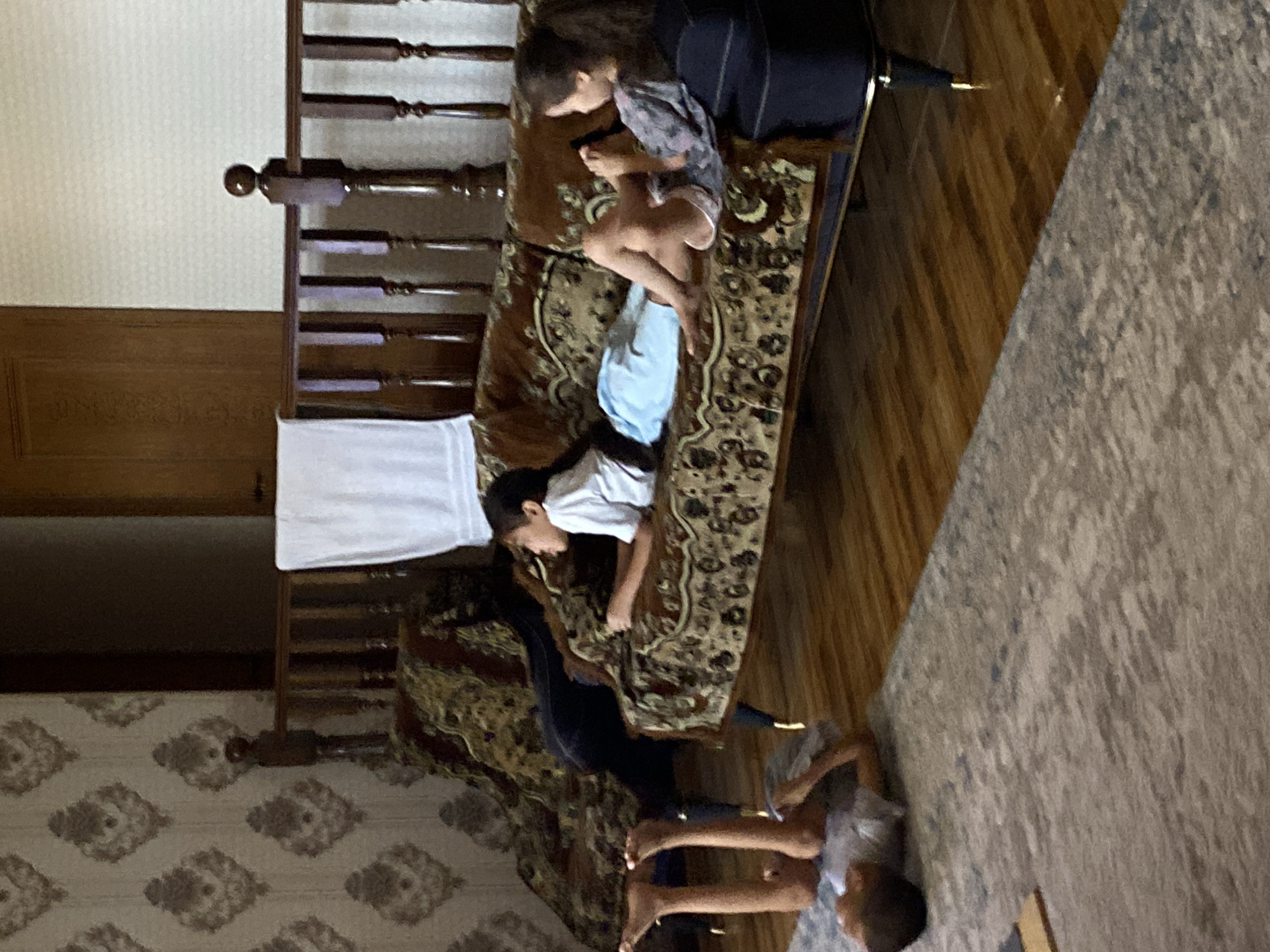 This image depicts an indoor scene, likely a living room, with a prominent vintage-style sofa. Three individuals are visible: two children are lying on a rug on the floor in the foreground, one in a yoga-like pose and the other lying on their back. A third child, a boy, is lying on the sofa, propped up on his elbow, wearing a white t-shirt and light blue shorts. Another child, a girl, is also on the sofa, to the right, looking at a phone and wearing a patterned top and shorts. The room has wooden floors and a patterned wallpaper. In the background, there is a wooden railing and a dark wooden door. The overall ambiance is casual and domestic. This scene is not identifiable as being in the city of Turkistan, Kazakhstan, based on visual cues alone.