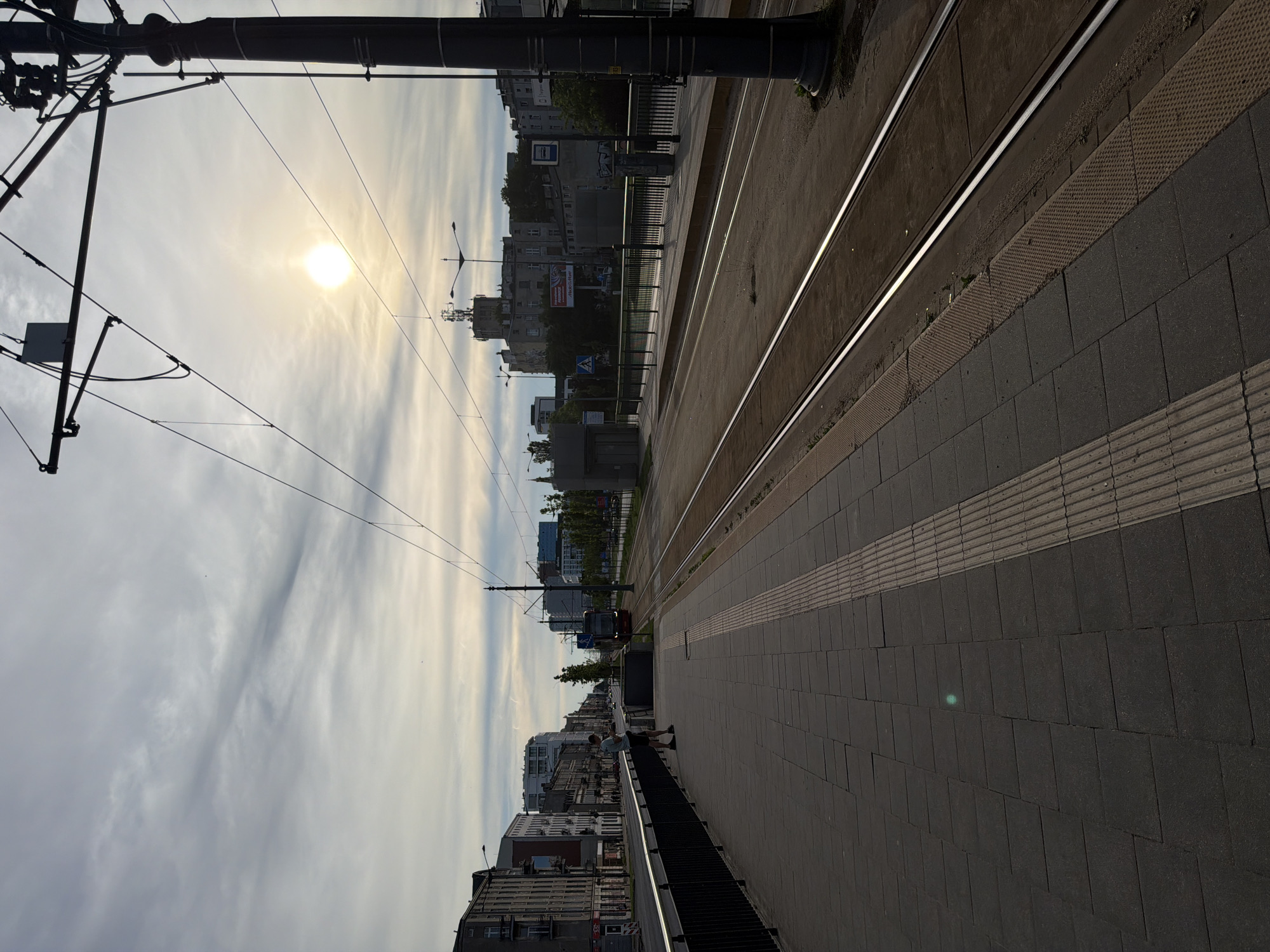The image depicts a street scene with a tram track and a sidewalk. The sun is visible in the sky, casting a warm glow. A person stands on the sidewalk, looking at their phone. A tram is approaching on the tracks. Buildings line the street, and there are trees and streetlights visible. The overall impression is that of an urban environment on a clear day.