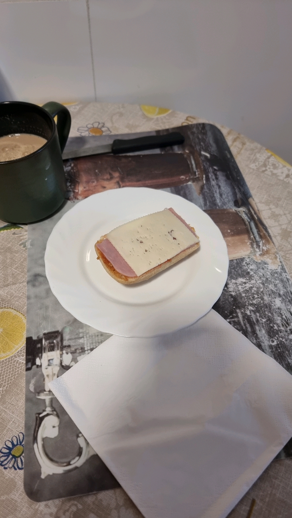 A close-up, slightly elevated shot shows a breakfast scene on a table. In the upper left corner is a dark green mug filled with a frothy, light-brown beverage, likely coffee or tea. To the right of the mug is a black-handled knife lying horizontally. In the center of the frame, a white plate holds a small, open-faced sandwich. The sandwich consists of a bread base topped with a spread, a slice of ham, and a slice of light-colored cheese with some perforations. The background features a patterned tablecloth or placemat with a yellow lemon slice and floral motifs. Beneath the plate and sandwich, a white napkin is folded. The overall impression is a simple, solitary breakfast setup in a domestic environment. There is no specific indication of the city of Sant Joan Les Fonts, Spain within the image itself.