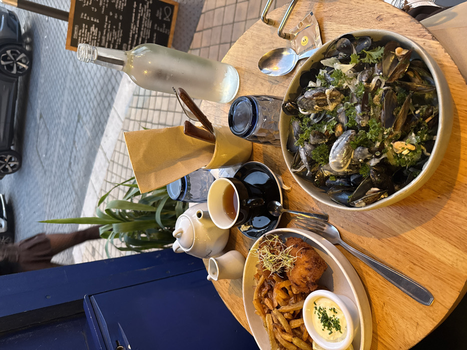 This image depicts an outdoor dining scene at a restaurant or cafe in Bordeaux, France, likely during the late afternoon or early evening.
In the foreground, a round, light-colored wooden table is set with a meal for two. On the left, an oval white plate holds a serving of golden-brown french fries next to two pieces of breaded, fried fish topped with green sprouts. A small white ramekin filled with a pale yellow sauce, garnished with chopped chives, is positioned on this plate. To the right of this plate, a silver fork rests on the table.
In the lower right, a large, off-white bowl contains a generous portion of mussels (moules) prepared in a creamy sauce and garnished with fresh chopped parsley. A silver spoon and a small white, square packet are situated beside this bowl.
Central to the table setup are a black ceramic teacup filled with dark liquid, resting on a matching saucer with a silver spoon, and a white ceramic teapot. Adjacent to the teapot is a small white ceramic creamer or milk jug. Two blue-tinted glasses, one upright and one inverted, are also present. A yellow ceramic holder contains several brown paper napkins and two brown wooden utensils, possibly spatulas or tongs. A frosted glass bottle, appearing to contain water, stands to the right of the yellow holder.
The background reveals a city street with grey rectangular paving blocks. A tall green plant is visible next to a dark blue door or wall section on the far left. Partially visible on the street are two cars: a light-colored sedan and a darker SUV further back. The lower body of a person, wearing a dark brown pleated skirt and dark patterned tights, is seen walking away from the camera near the blue wall. On the right, a wooden blackboard sign, supported by a wooden post, displays a menu with white writing, indicating typical restaurant offerings.