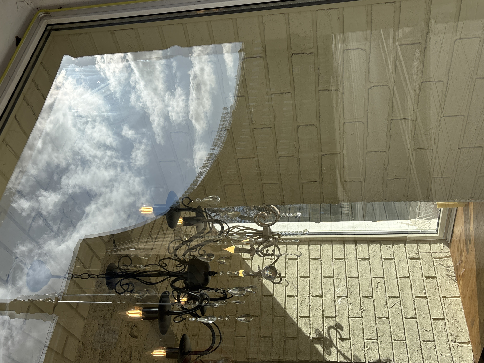 The scene depicts an interior space viewed through a large window, which simultaneously reflects the exterior environment. In the interior, a dark, ornate metal chandelier with multiple arms is centrally positioned, featuring electric candle-style bulbs emitting a warm yellow light. Numerous clear, faceted crystal pendants adorn the chandelier. A similar, smaller light fixture or a portion of another chandelier is visible on the right. The interior walls are composed of light-colored, textured brickwork, likely painted a cream or pale yellow hue, with strong diagonal shadows from the chandelier and window frame cast upon them by direct sunlight. A polished wooden surface, likely a windowsill or countertop, is visible at the bottom of the frame, also illuminated by sunlight.
Reflected on the window glass is a bright, partially cloudy sky, exhibiting patches of white cumulus clouds against a blue background. Beneath the sky reflection, part of a light-colored, possibly concrete or stone, building with a flat roofline and a prominent upper edge or parapet is discernible. No individuals are present in the image. The location is Saint Petersburg, Russia.