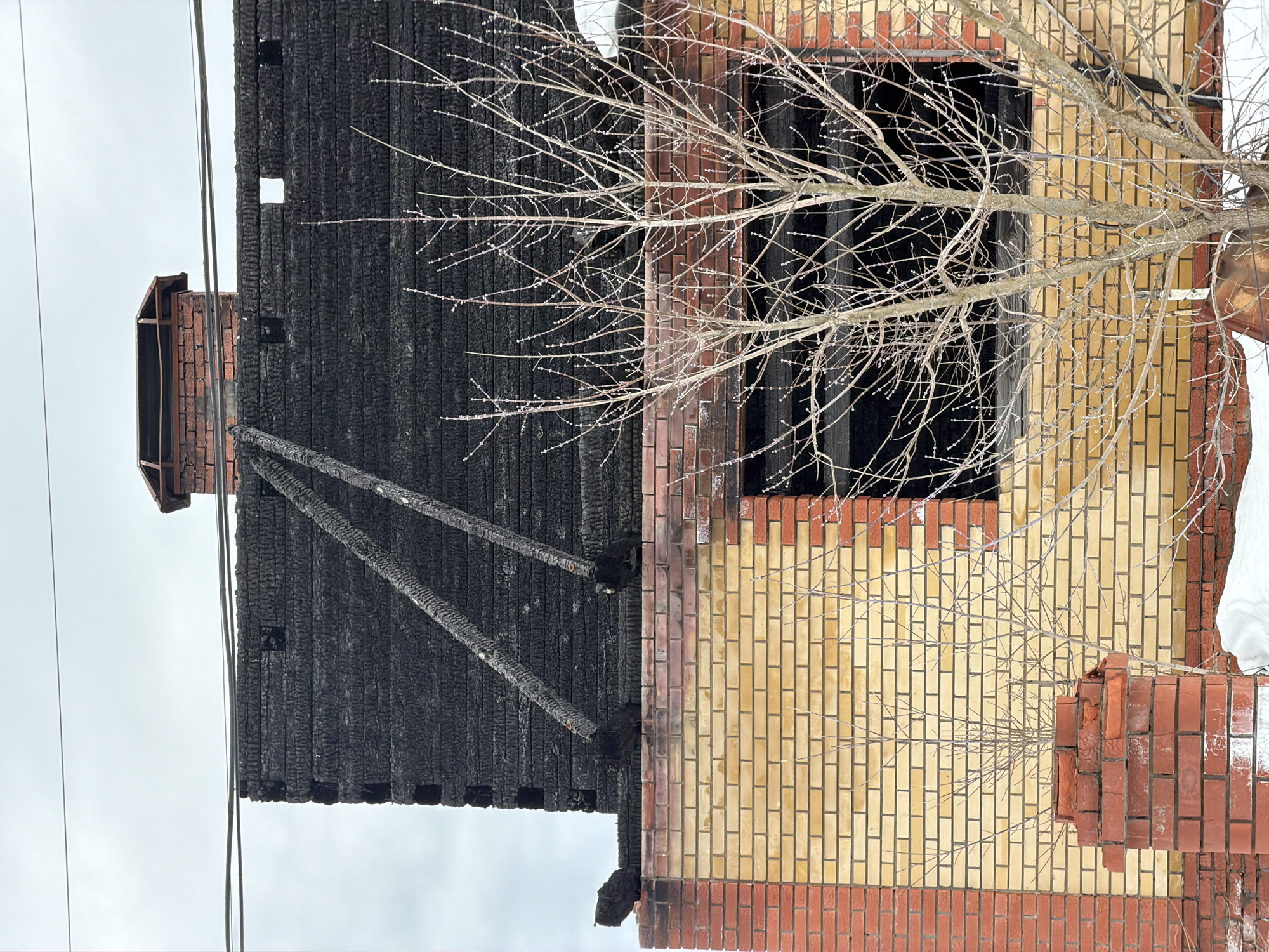The image depicts a two-story residential structure in Chaikovsky, Russia, exhibiting extensive fire damage, likely captured during winter or early spring. The lower level is constructed of light yellow brick with red brick corner sections, displaying distinct soot and heat discoloration along its upper perimeter. The entire upper level and roof structure are severely charred and blackened, appearing to be of timber or log construction, with individual burned horizontal members clearly visible. A red brick chimney with a damaged metal cap protrudes from the blackened roofline on the upper left, partially obscured by two horizontal power lines. Two prominent, diagonally oriented charred wooden beams are affixed to or protrude from the blackened upper wall. A large, empty window opening on the first floor's right side provides a view into the dark interior. In the foreground, the ground is covered with snow, and several leafless deciduous trees or shrubs with nascent buds stand, partially obscuring the building's facade. No individuals are present in the scene. The sky is overcast and grey.
