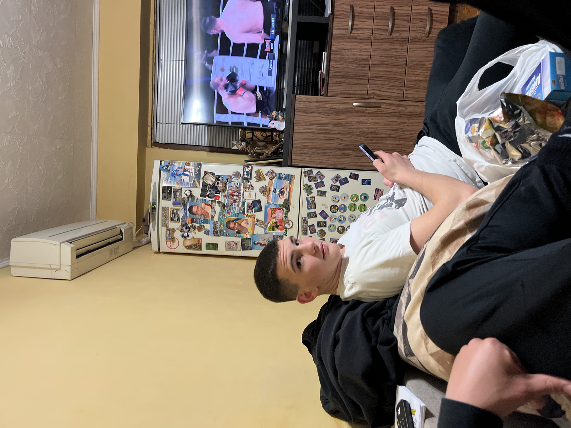 A young man is reclining on a dark-colored couch, holding a mobile phone in his right hand. He is looking towards the camera. Next to him, there is a white plastic bag filled with snacks and a blue box. A TV is showing a boxing match. The background includes a refrigerator covered with magnets and a storage unit. The scene appears to be inside a residential building, with light beige-colored walls. There is no specific indication of this image being from Burgas, Bulgaria.