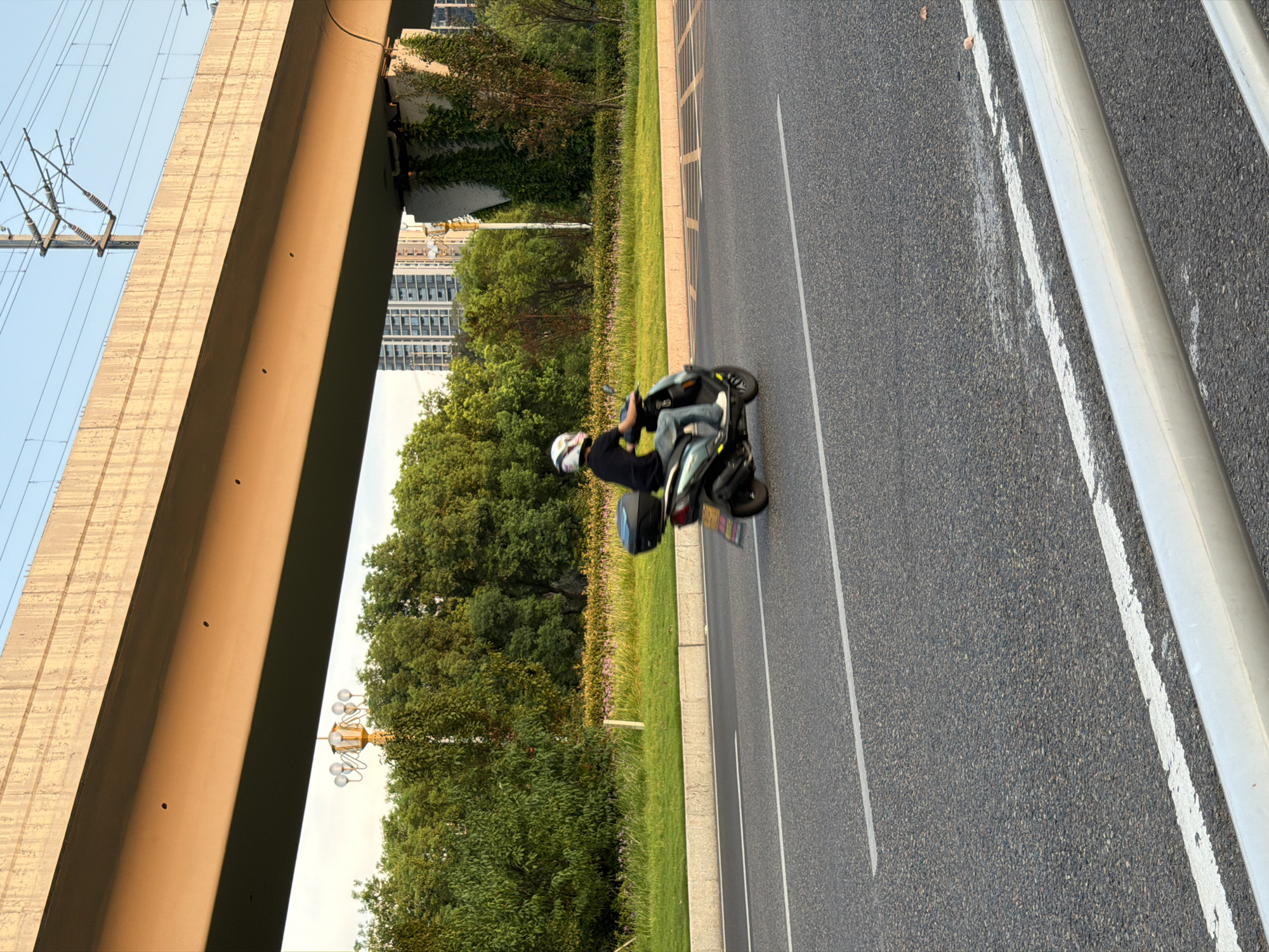 A person on a scooter rides along a multi-lane asphalt road. The rider wears a black shirt, blue jeans, and a white helmet. The scooter is dark colored with a black storage box on the back. The road has white lane markings. In the background, a large concrete overpass, possibly for a highway or train, spans across the scene. Beyond the overpass, there are green trees, some buildings, and overhead power lines. A street lamp with multiple globes is visible on the left side of the road, near the trees. The lighting suggests it is either early morning or late afternoon. The location is Jiangjin District, Chongqing, China.