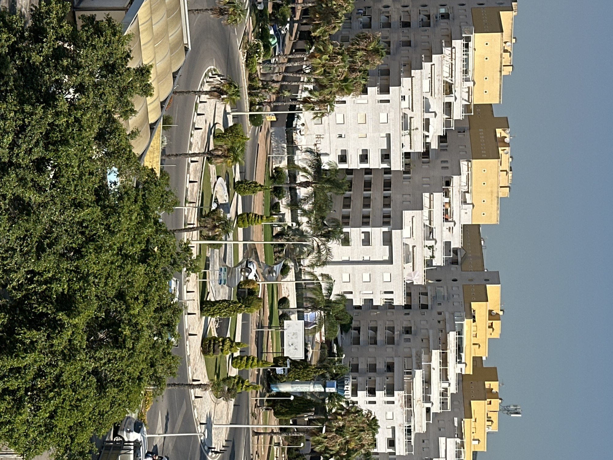 This image captures an aerial view of a modern urban landscape, featuring a roundabout at the intersection of several roads. Dominating the scene are large, multi-story apartment buildings with a distinctive architectural style, characterized by a lighter colored base and bright yellow upper sections. The buildings have numerous balconies and windows, indicating residential occupancy. The roundabout itself is landscaped with manicured bushes, palm trees, and conical shaped trees, surrounding a central fountain. Several cars are visible on the roads, indicating traffic flow. Streetlights are positioned along the roadside. In the background, the sky is clear and blue, suggesting a sunny day. The scene appears to be located in a well-developed area with contemporary infrastructure and landscaping.