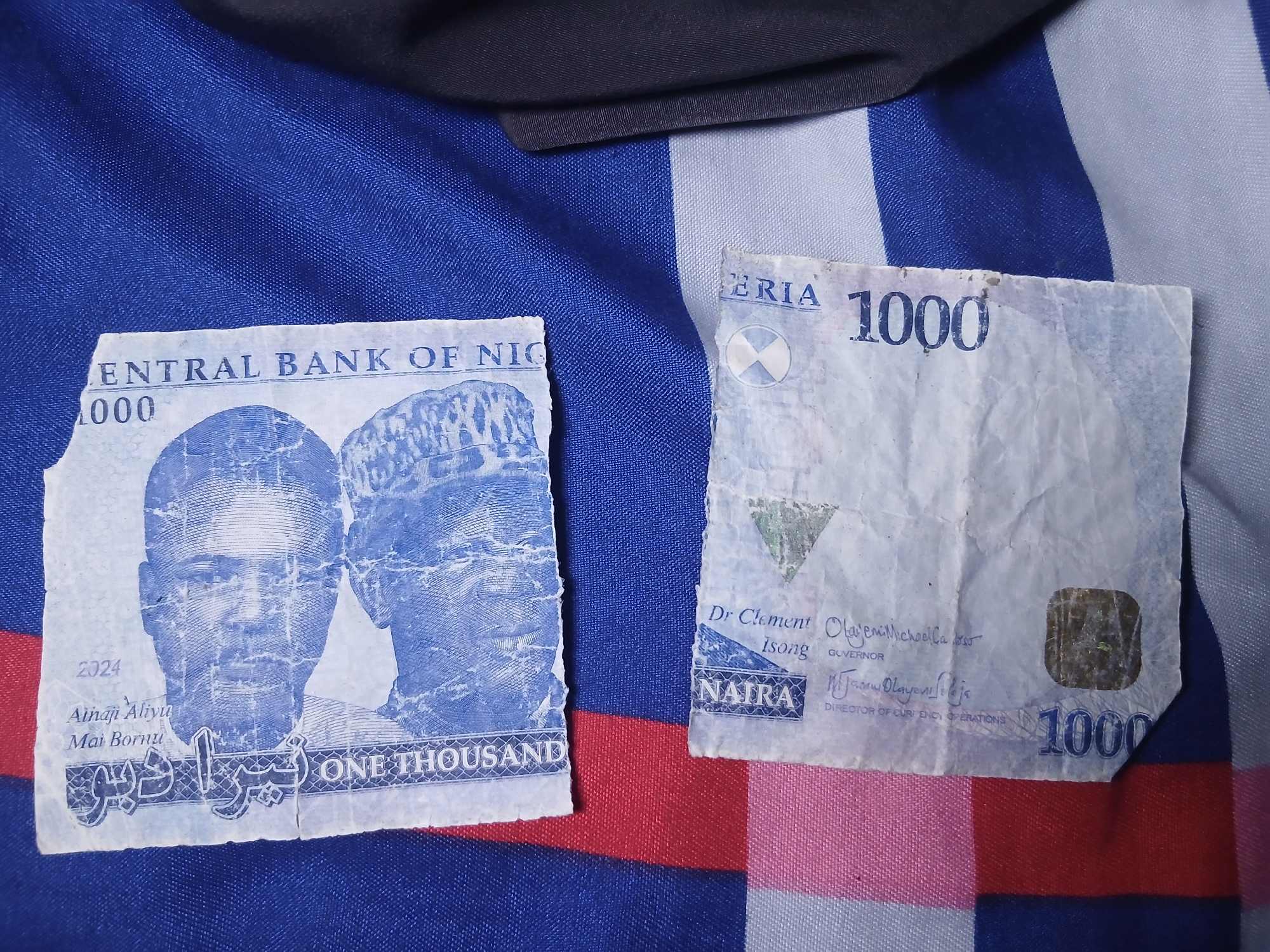 The image shows two torn halves of a Nigerian 1000 Naira banknote. The left piece features the portraits of Alhaji Aliyu Mai Bornu and another man, along with the words "CENTRAL BANK OF NIG" and "ONE THOUSAND." The right piece displays "ERIA 1000," "NAIRA," and signatures of Dr. Clement Olajemi Michael Isong, Governor, and a signature denoting the Director of Currency Operations. The banknote pieces are placed on a surface with blue, white, and red stripes, with a dark fabric partially visible in the background. There is no information in the image to determine a location in the city of Lagos, Nigeria.