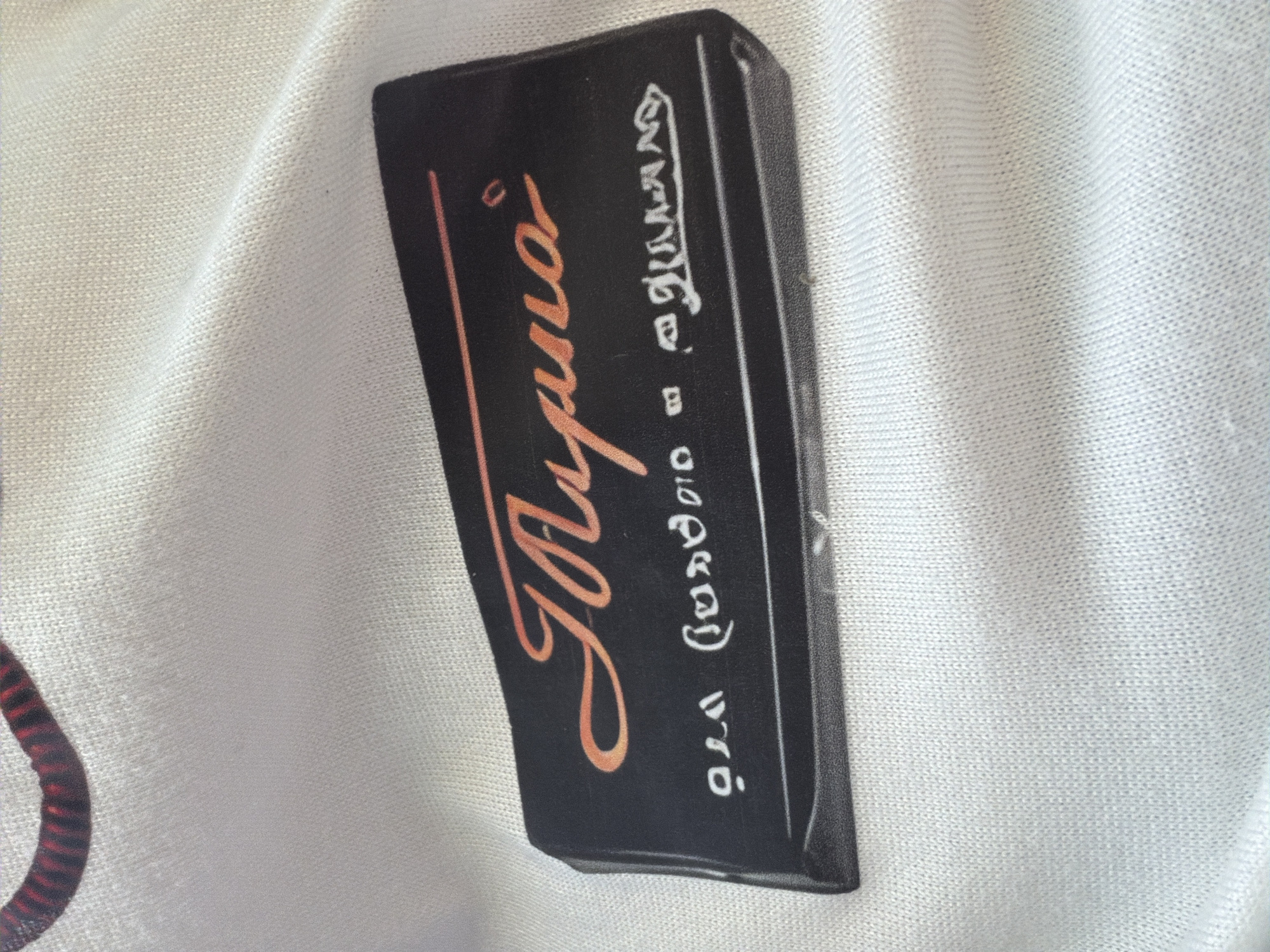 A close-up, high-angle shot shows a black rectangular label with an orange script logo and white text on a white knitted fabric. The logo appears to read "Tusna" in an elegant cursive font. Below the logo, white text is visible, with symbols that resemble Cyrillic letters. The fabric is creased and textured, suggesting it is clothing. The scene does not contain any specific elements that place it in Sochi, Russia.