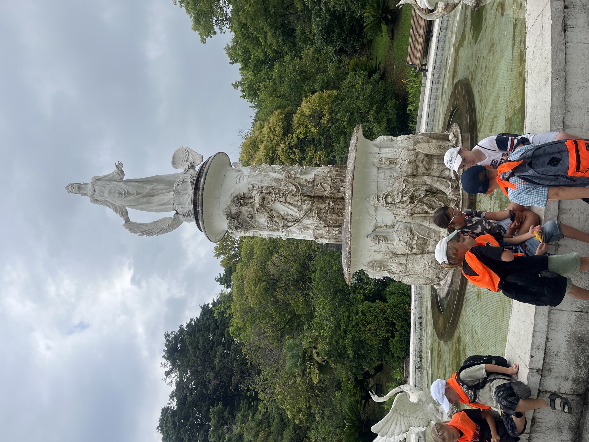 The image shows a fountain adorned with sculptures, featuring a statue of a woman at the top, winged figures, and bas-reliefs of various figures along its tiers. The fountain is situated in a park-like setting with lush green trees and foliage in the background. In the foreground, several children are seated on the edge of the fountain. They are wearing orange vests and backpacks, suggesting they are part of a tour group or school outing. Some of the children are interacting, looking at phones or talking to each other. The sky is overcast, indicating a cloudy day. This scene is likely in the city of Sochi, Russia, known for its numerous parks and ornate fountains, often associated with its history as a resort town.