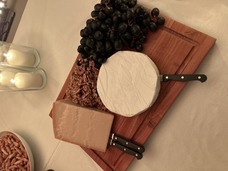 A wooden cutting board displays a selection of food. A bunch of black grapes sits atop the board. There is a pile of walnuts, a round of brie, and a block of hard cheese. Two black-handled knives are present, one piercing the brie, the other resting near the hard cheese. A bowl of nuts and candle-filled glass bottles are also visible on the white surface surrounding the board.
