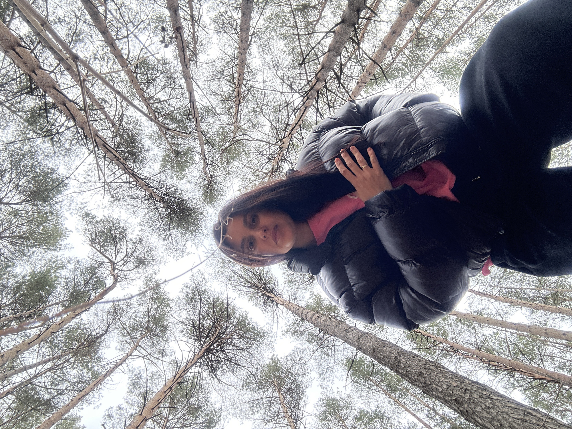 A low-angle photograph depicts a woman standing in a forest near Chaikovsky, Russia. She is wearing a black puffer jacket over a pink hooded sweatshirt. She has long, dark hair and is looking down at the camera, with her right hand raised near her face. In the background, tall, slender trees reach upwards, their branches forming a dense canopy overhead. The sky is overcast.